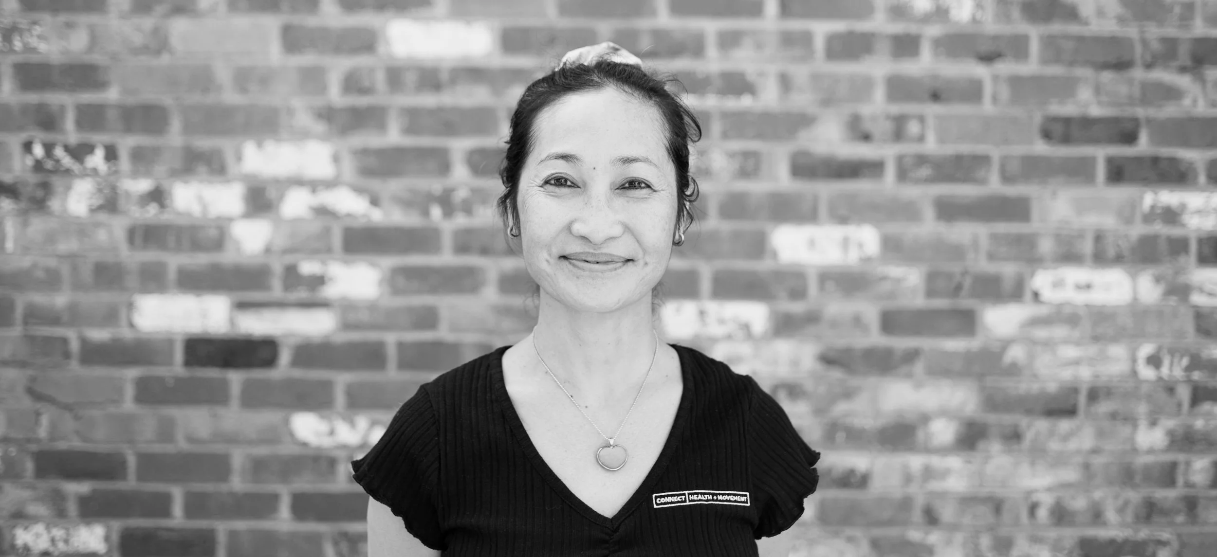 A woman with dark hair tied back, wearing a black top with a badge reading "Connect Health + Movement," standing in front of a brick wall.