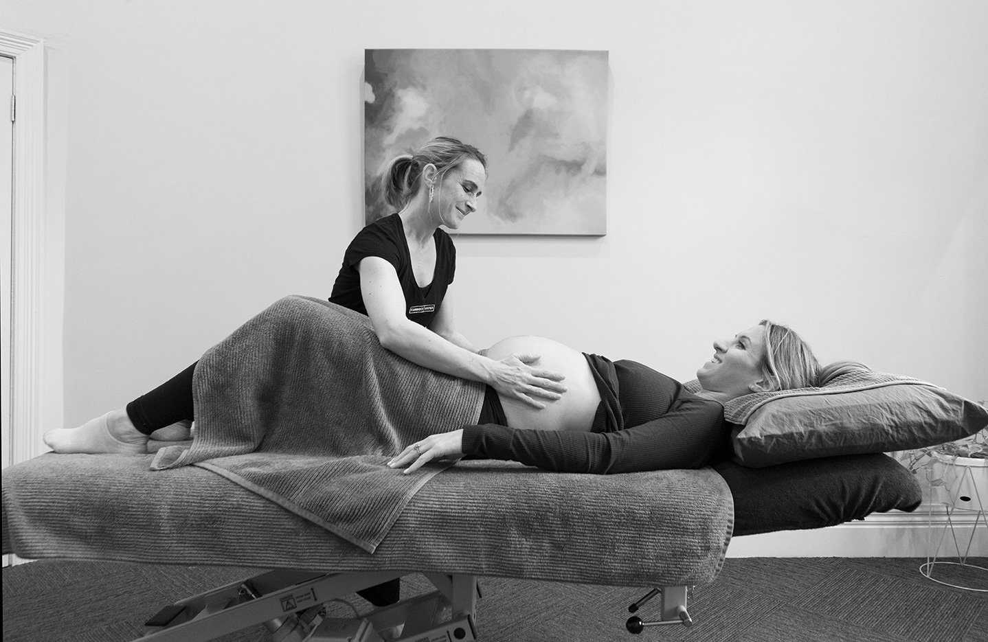 A woman lying on a medical examination table with her head on a pillow, smiling, as a healthcare professional checks her pregnant belly in a clinical setting.