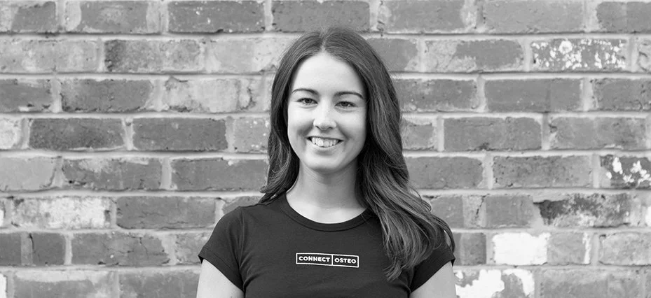 Black and white photo of a young woman with long wavy hair smiling, standing in front of a brick wall, wearing a T-shirt with the words "CONNECT OSTEo" on it.