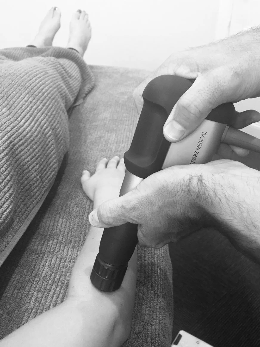 A medical professional uses an ultrasound device on a person's forearm. The person is lying on a couch with their legs stretched out and is wearing a knitted pair of pants.