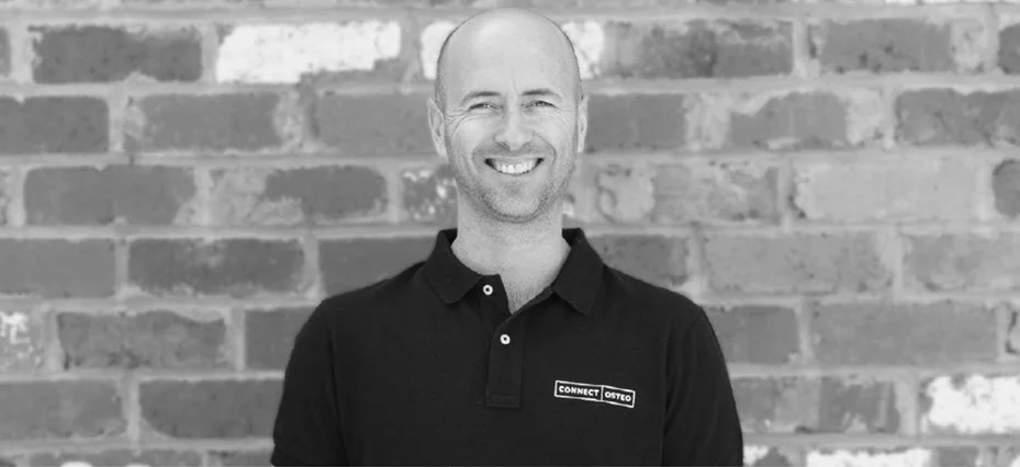 A man with a bald head and a beard smiling, standing against a brick wall, wearing a black collared shirt with a name tag.