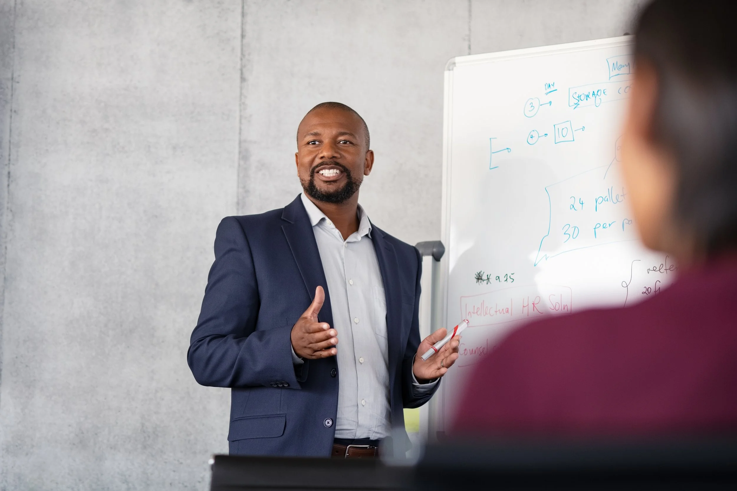 A man in a dark blue suit and light blue shirt standing in front of a whiteboard, speaking to a group in a modern office setting.