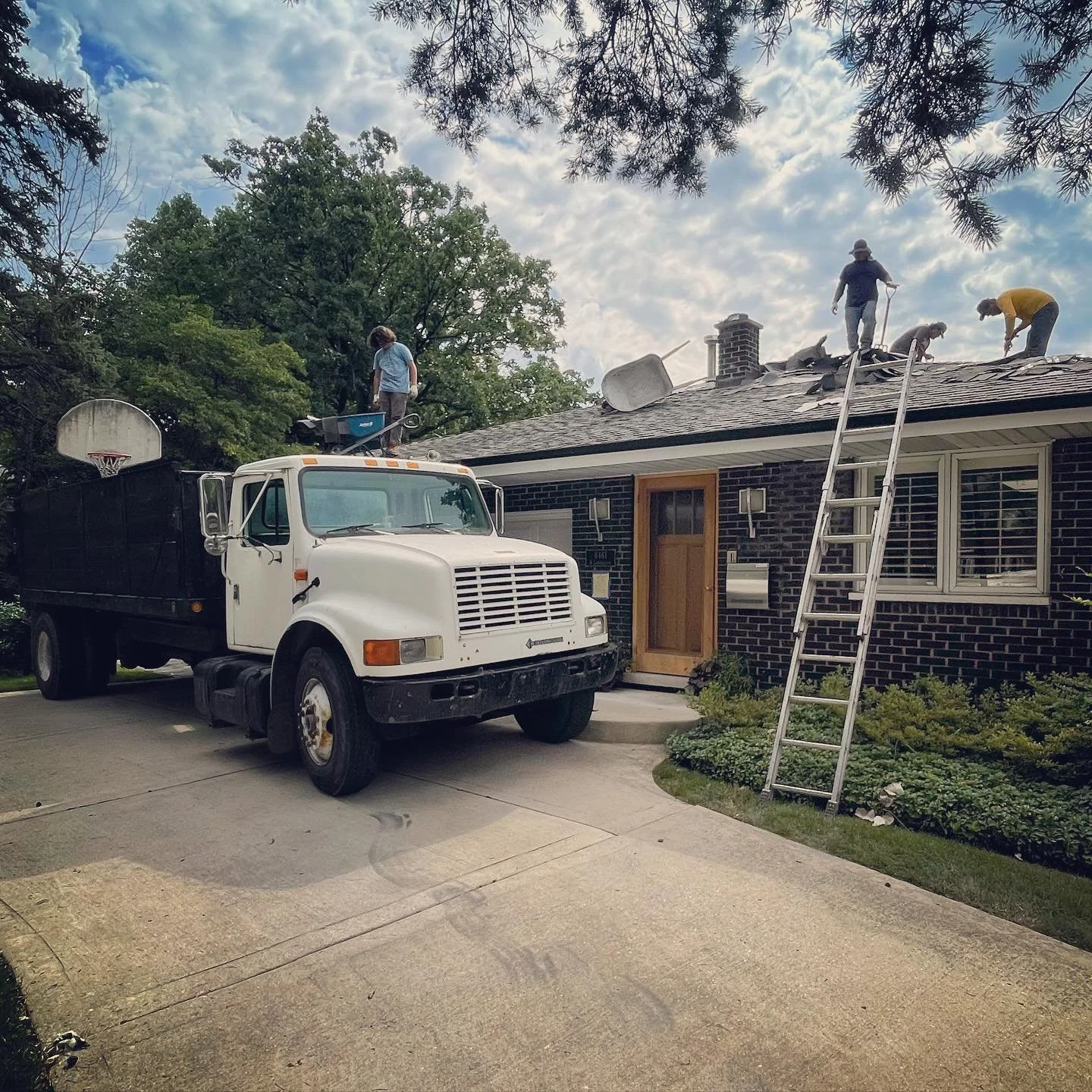 People working on a house roof with a ladder, a truck parked in the driveway, and a basketball hoop in the yard.