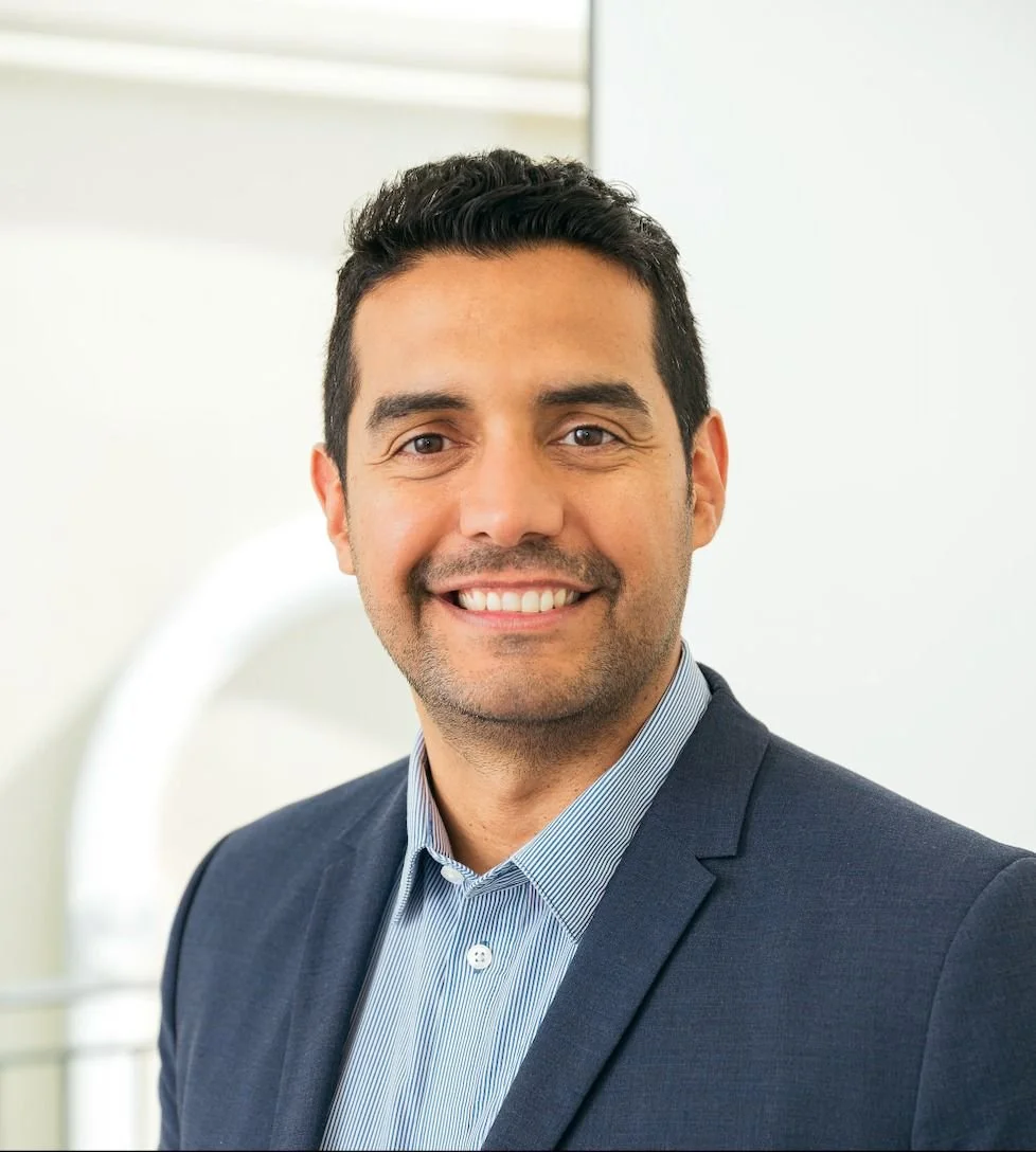 Professional headshot of a smiling man in a suit and dress shirt, standing indoors with a blurred background.