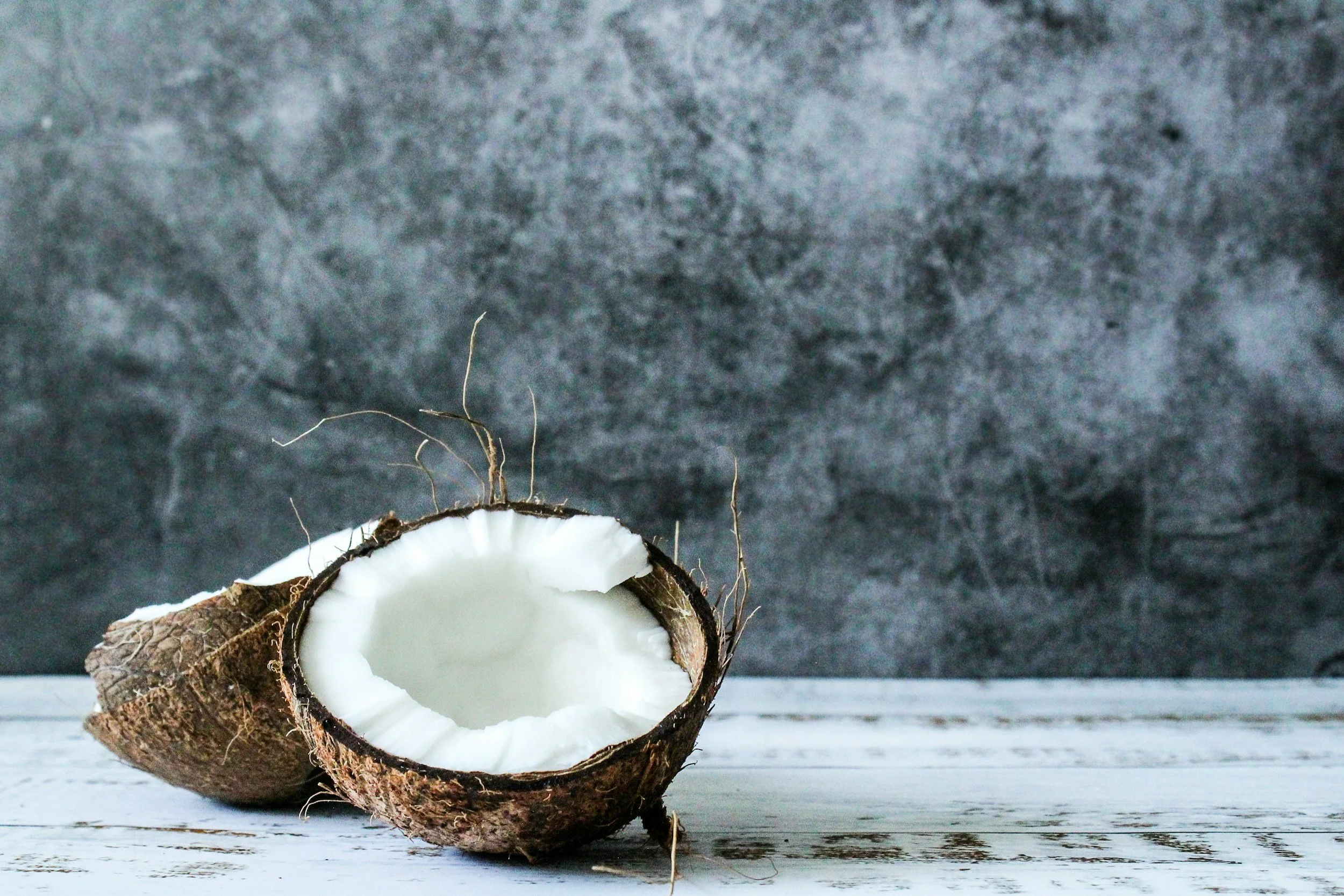 Half of a coconut with white flesh and rough brown outer shell, placed on a white wooden surface against a dark gray textured background.
