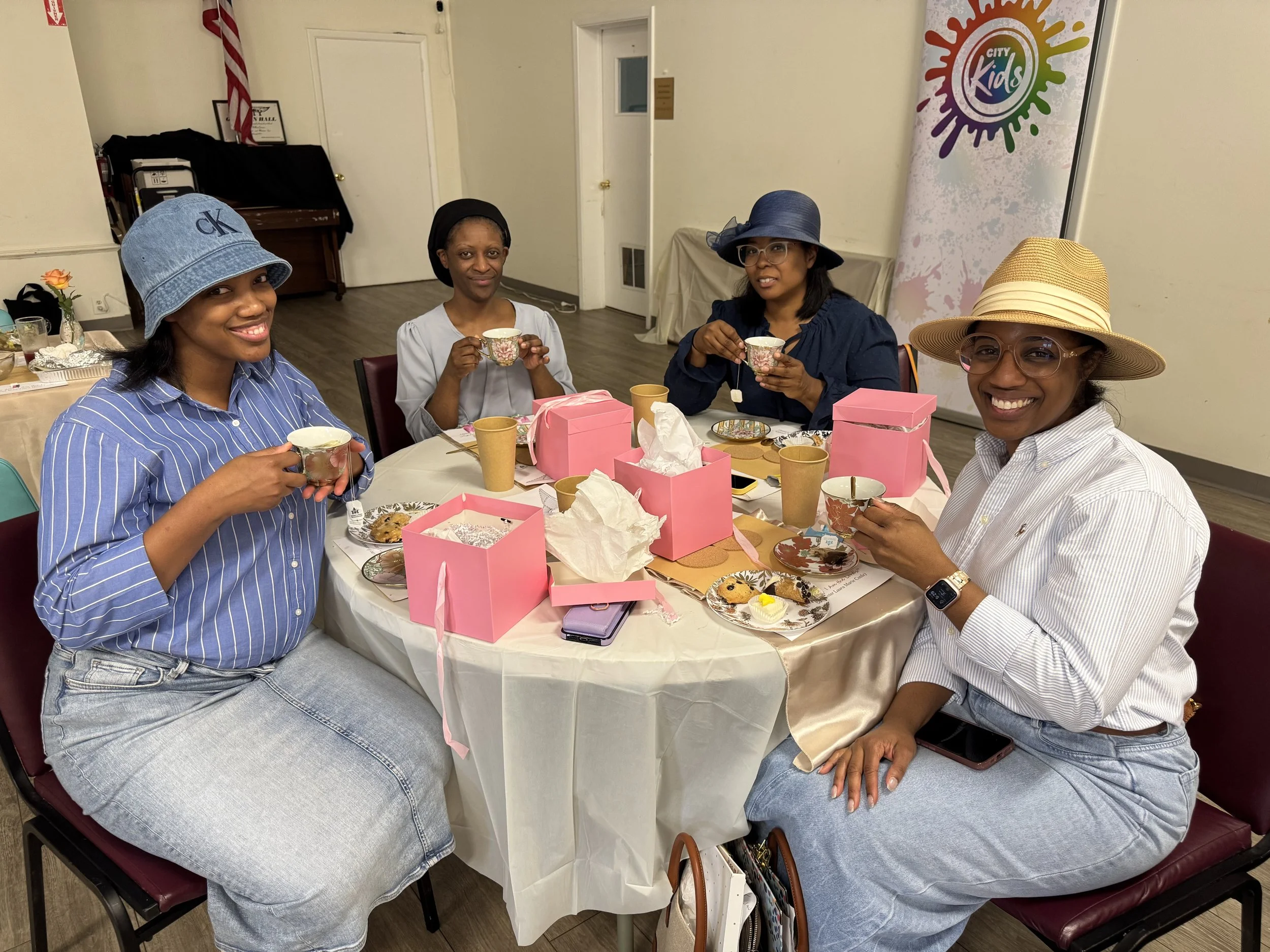 Four women sitting around a table with pink gift boxes, tea cups, and desserts, smiling and enjoying tea together at a celebration event.