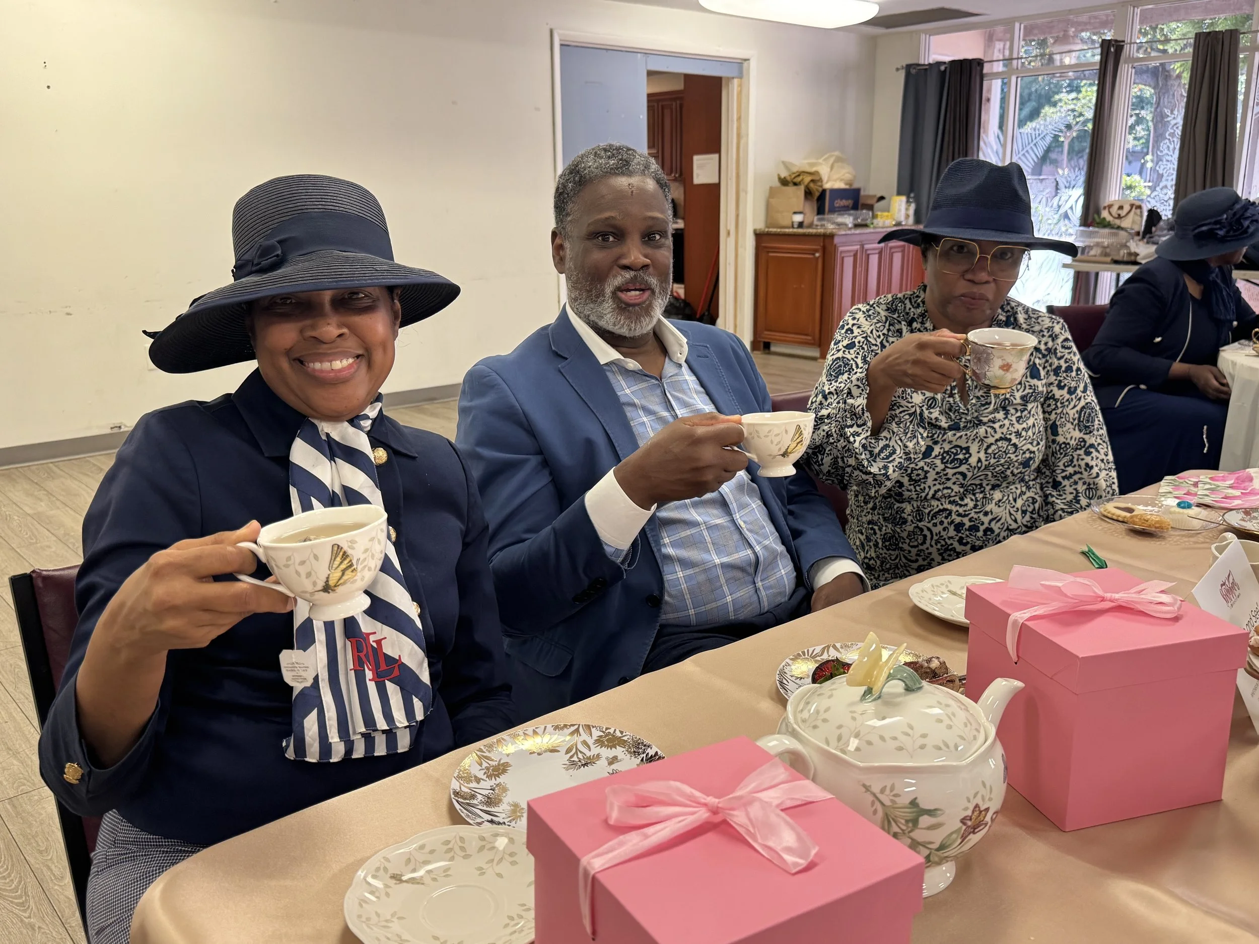 Sis. Angela Hughes, Bishop Anthonee Patterson, and Sis. Vivian Shelton at a table drinking tea, smiling, with pink gift boxes and tea sets on the table, in a decorated room with large windows.