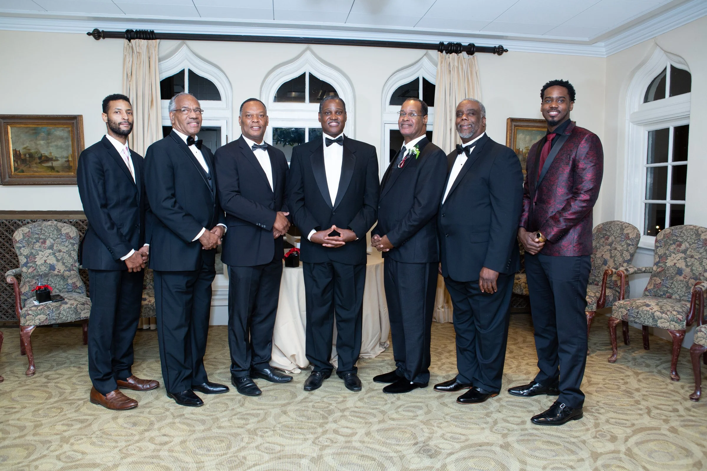 Group of eight men of the presbytery dressed in tuxedos standing in a formal room with large windows and artwork
