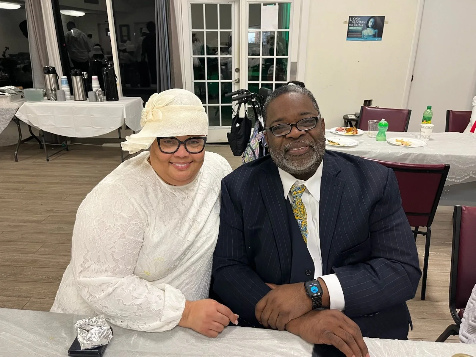 A woman dressed in white with glasses and a white hat sitting next to a man in a suit with glasses and a smartwatch, smiling at a social gathering or celebration.