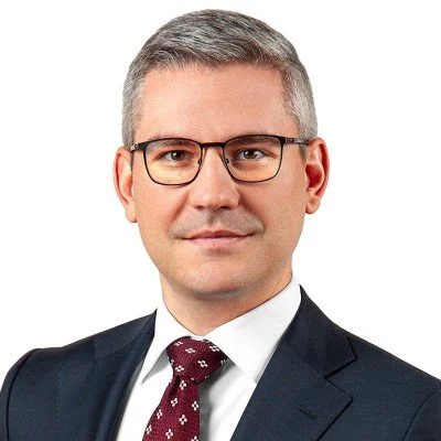 Portrait of a man with gray hair and glasses wearing a dark suit, white shirt, and red patterned tie.