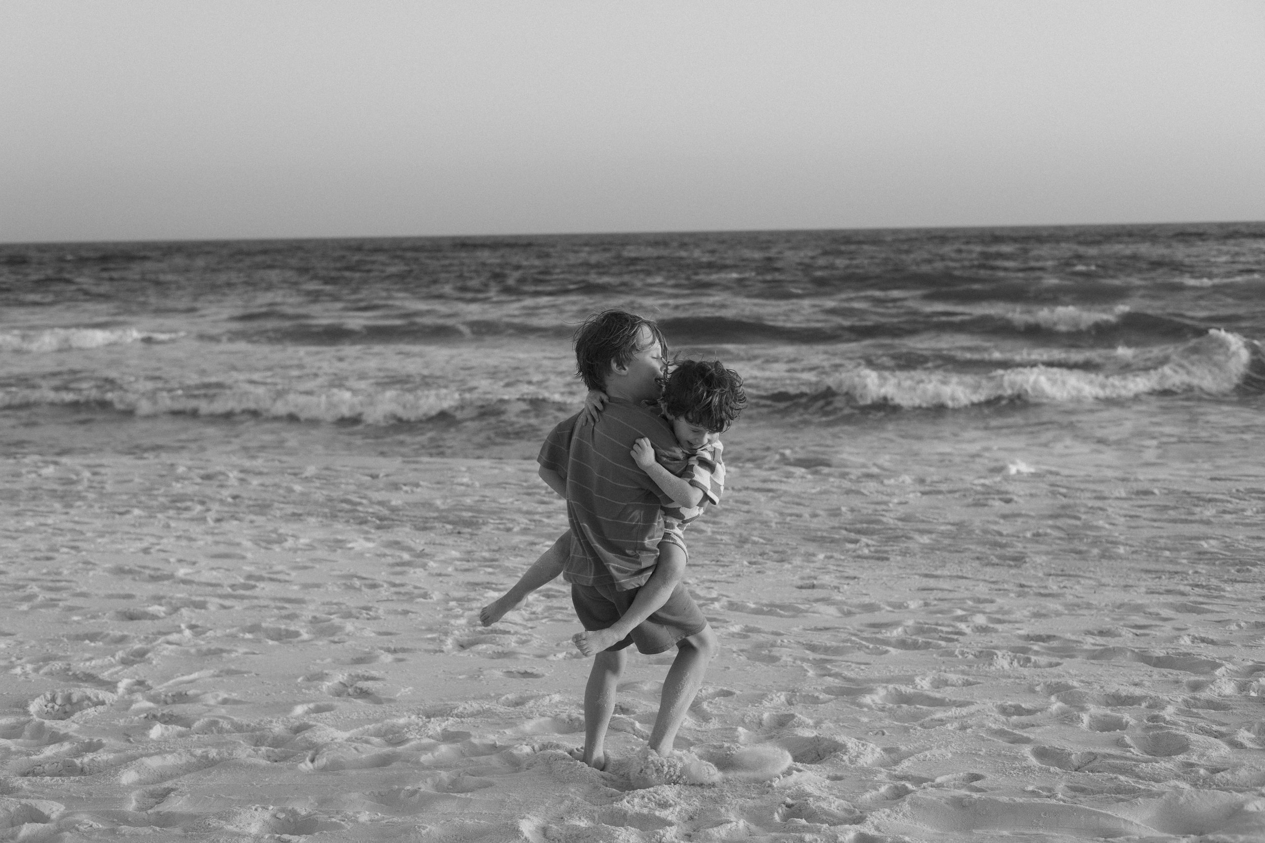 Two children playing on the beach, one carrying the other, with waves in the background.