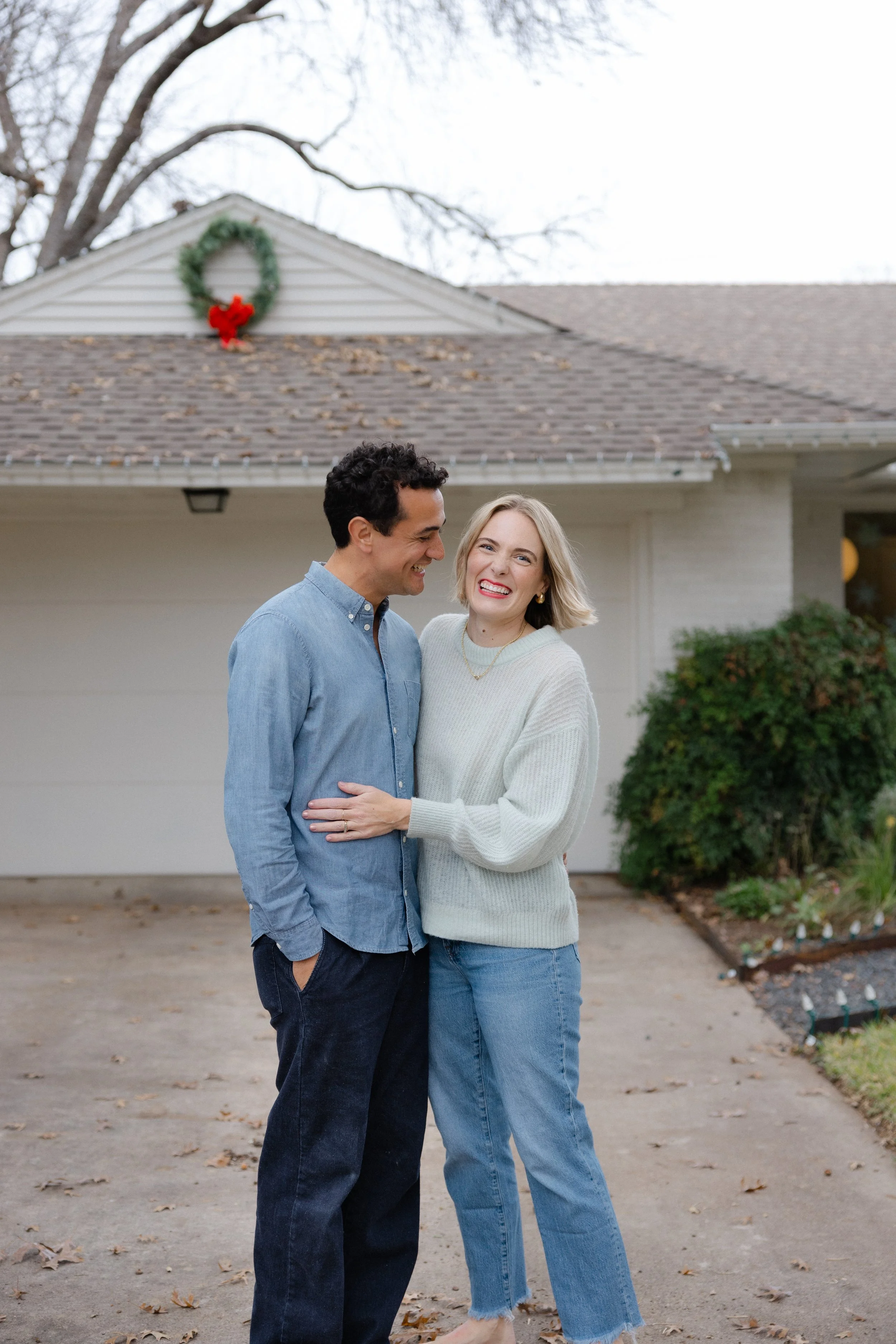 A smiling couple stands in front of a house decorated for the holidays with a wreath and red bow on the garage.