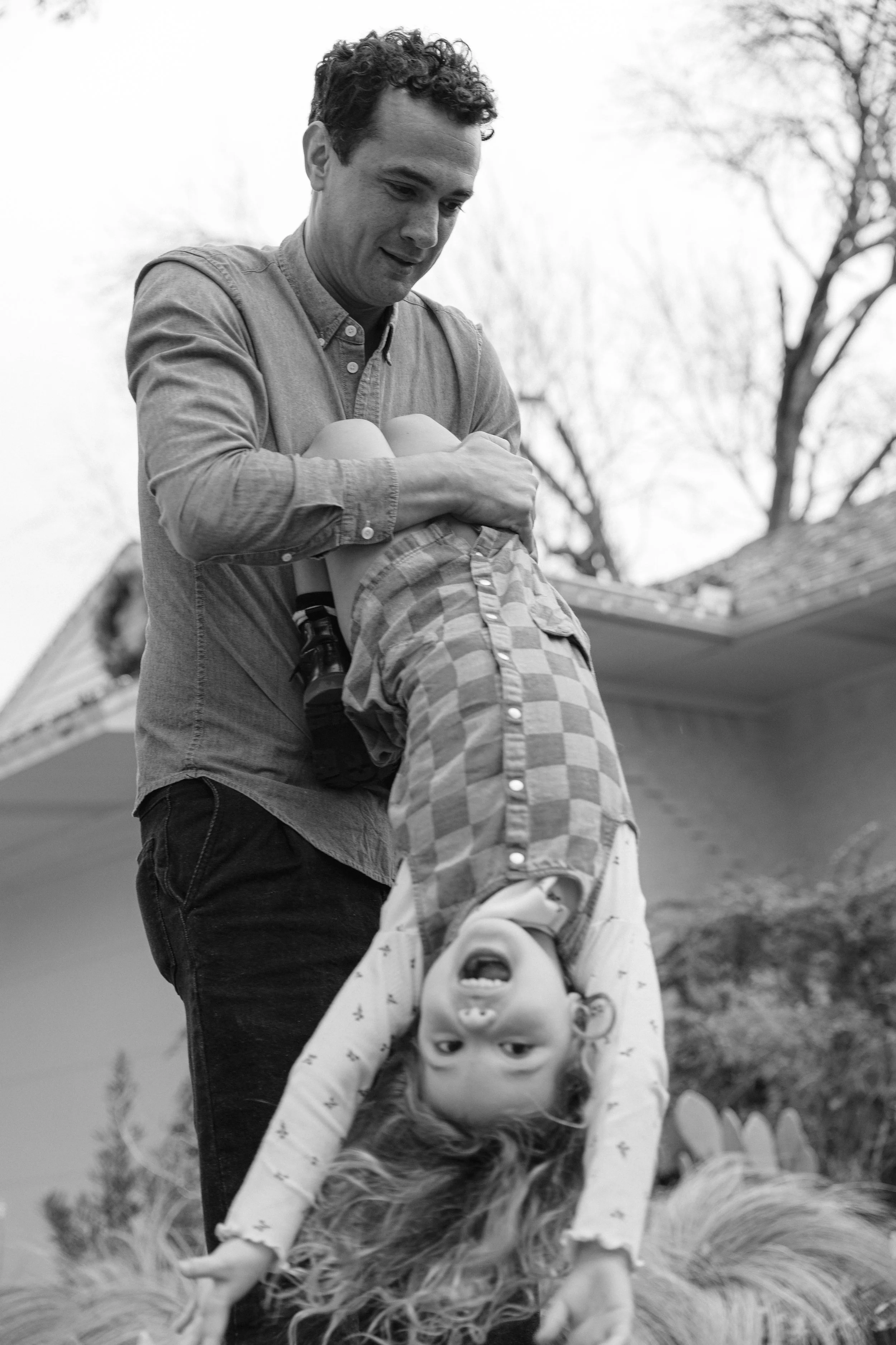 A man holding and swinging a young girl upside down outside near a house, with trees in the background, black and white photo.