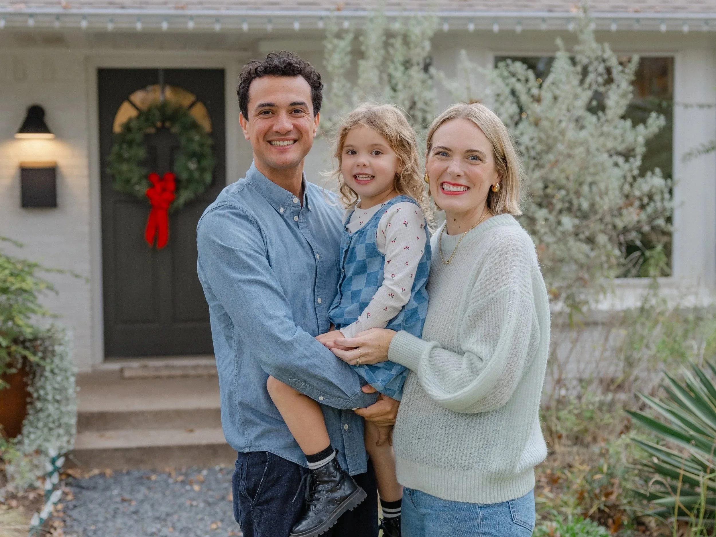 A happy family of three standing outside their home, smiling at the camera. The father is holding a young girl, and the mother is standing beside them. There is a Christmas wreath on the door in the background.