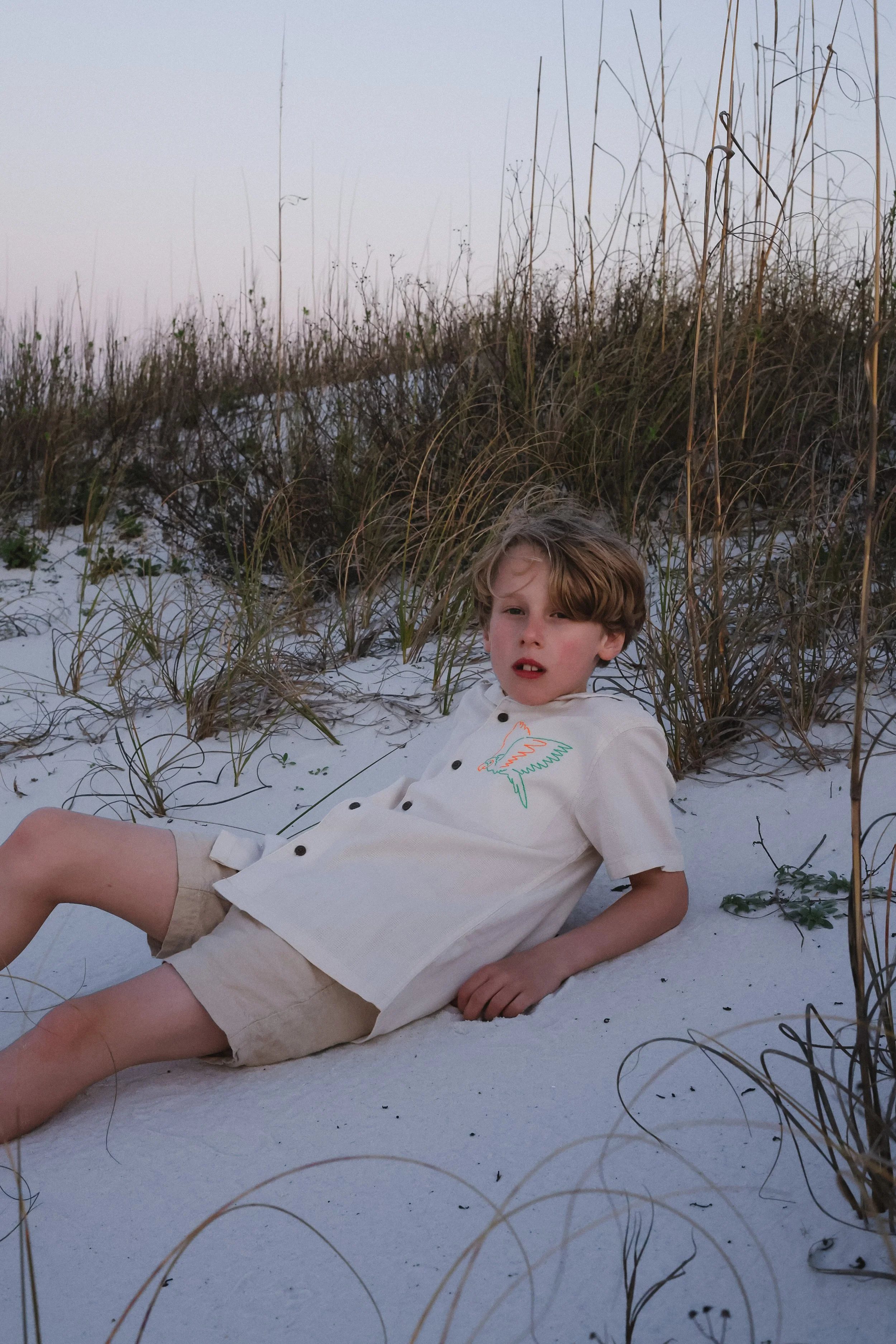 A young boy lying on white sand at the beach, surrounded by tall beach grass, during sunset or dusk.