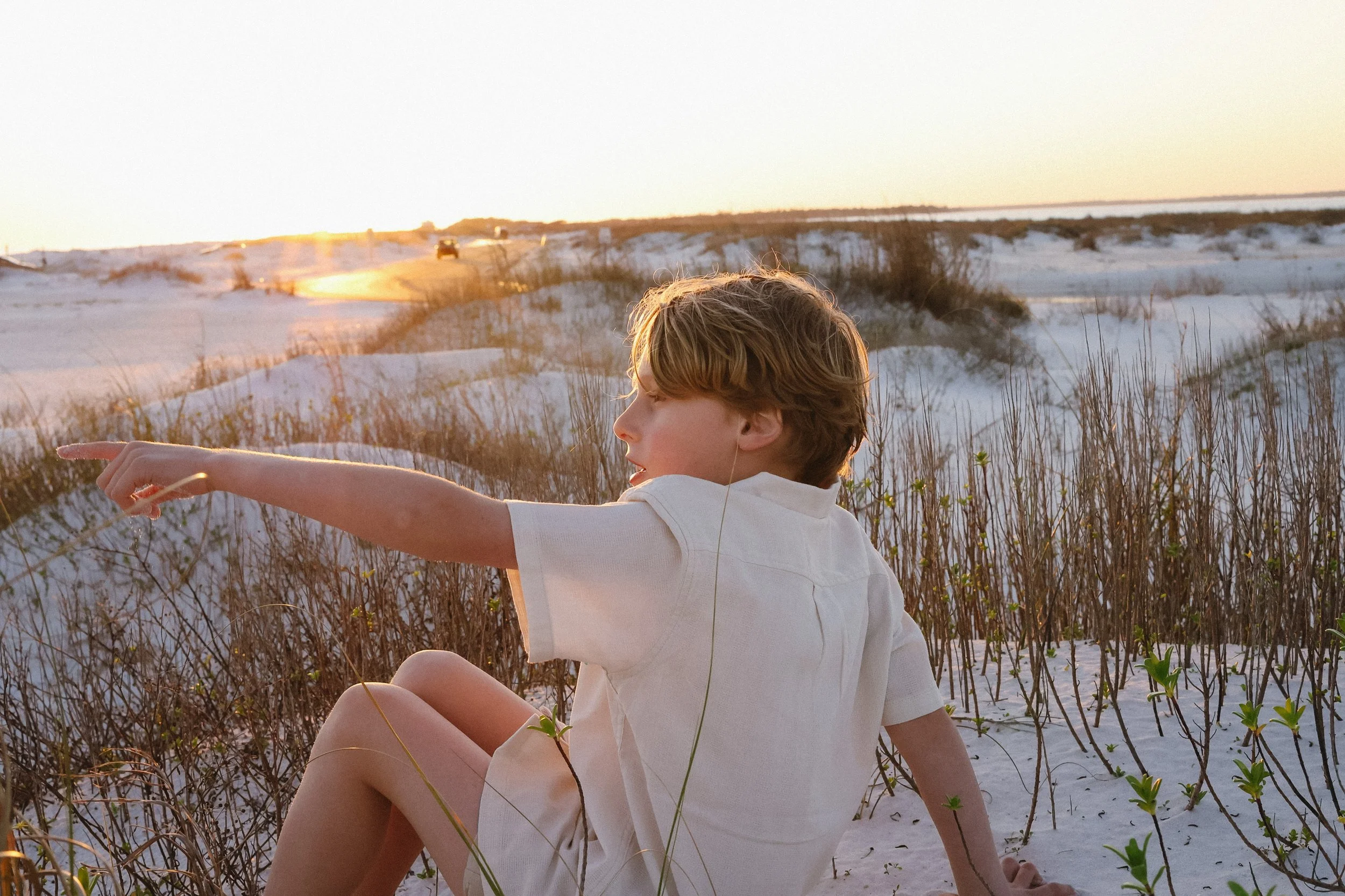 A young boy with blond hair wearing a white shirt and shorts sitting on sandy dunes with sparse vegetation during sunset, pointing into the distance.