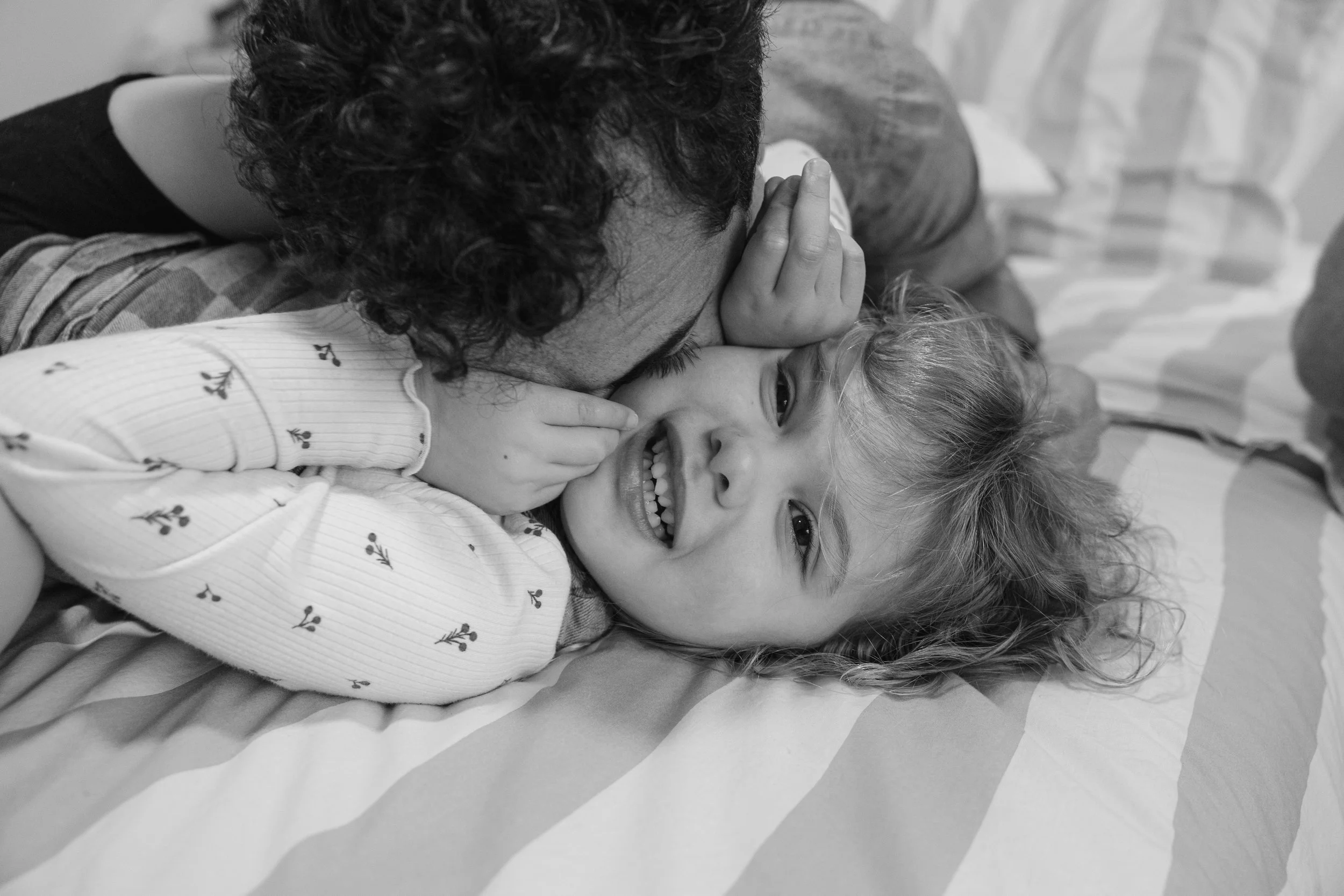 Father and daughter, one with curly hair and another with straight hair, are playfully hugging and smiling on a bed as one kisses the other's forehead in a joyful moment.