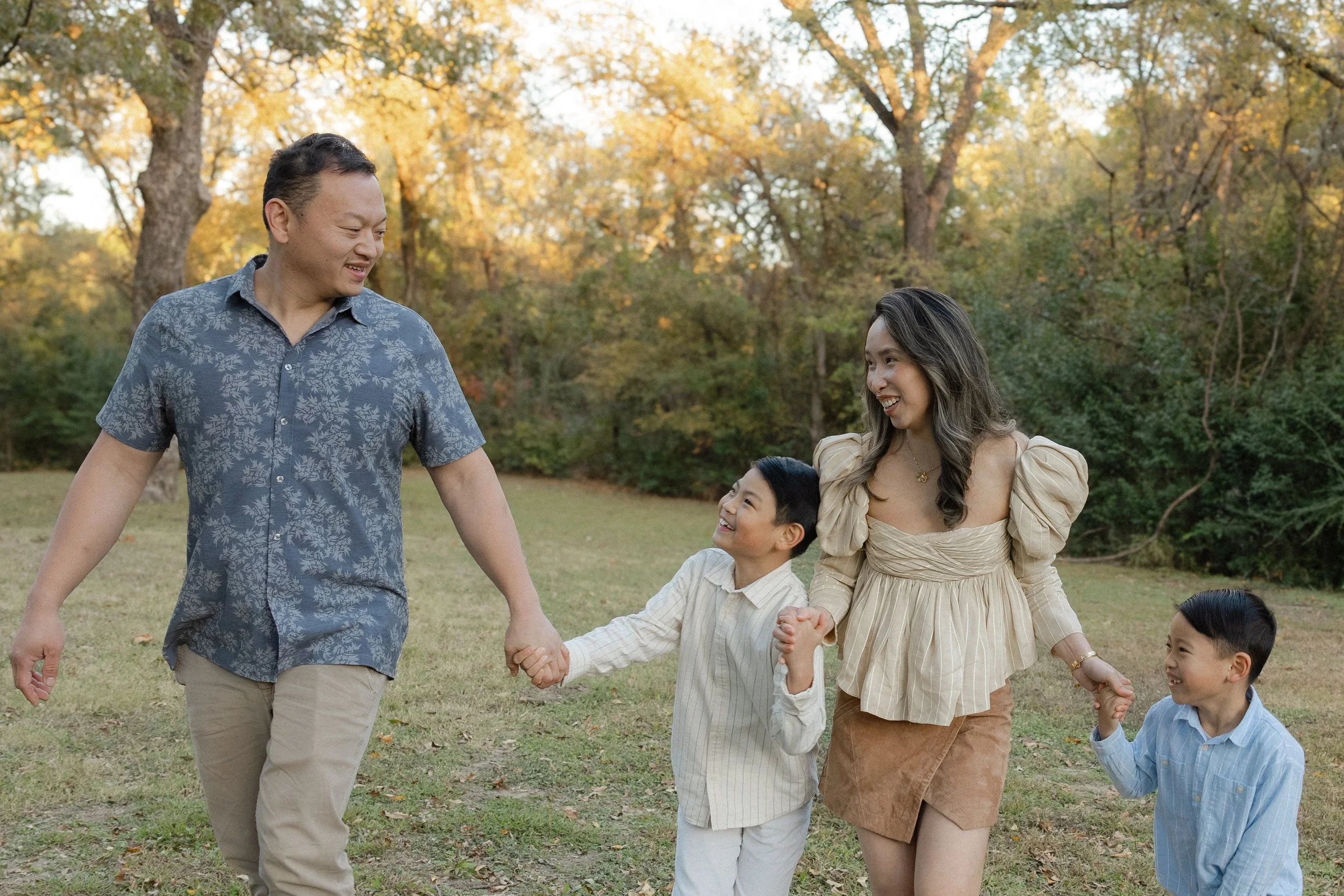 A family of four, including two adults and two children, holding hands and walking together outdoors in a park during fall, smiling and enjoying a sunny day.