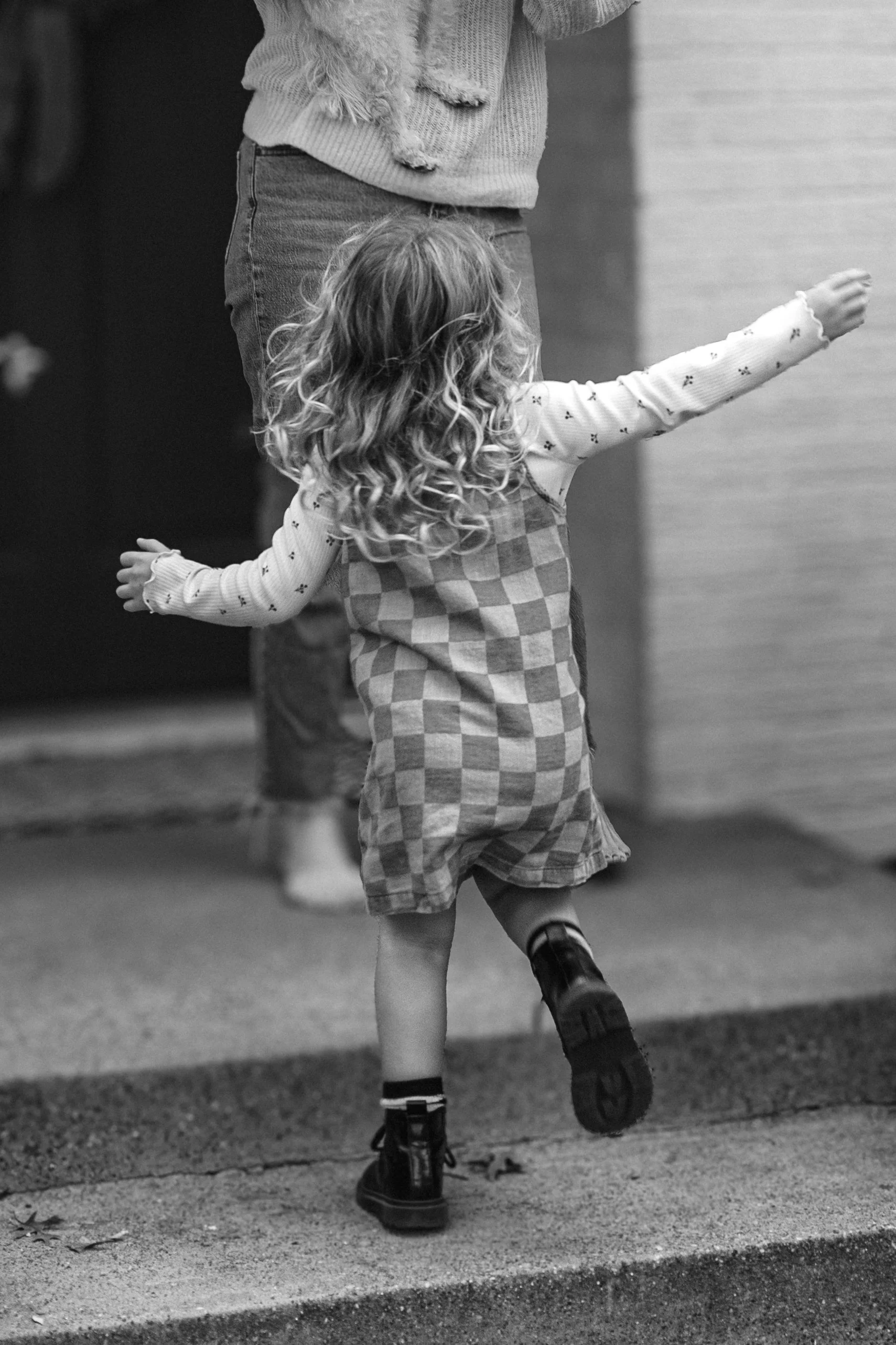 A young girl with curly hair, wearing a checkered dress and boots, is walking or running on the sidewalk with her arms outstretched, in front of an adult whose torso and legs are partially visible. childhood moments captured.