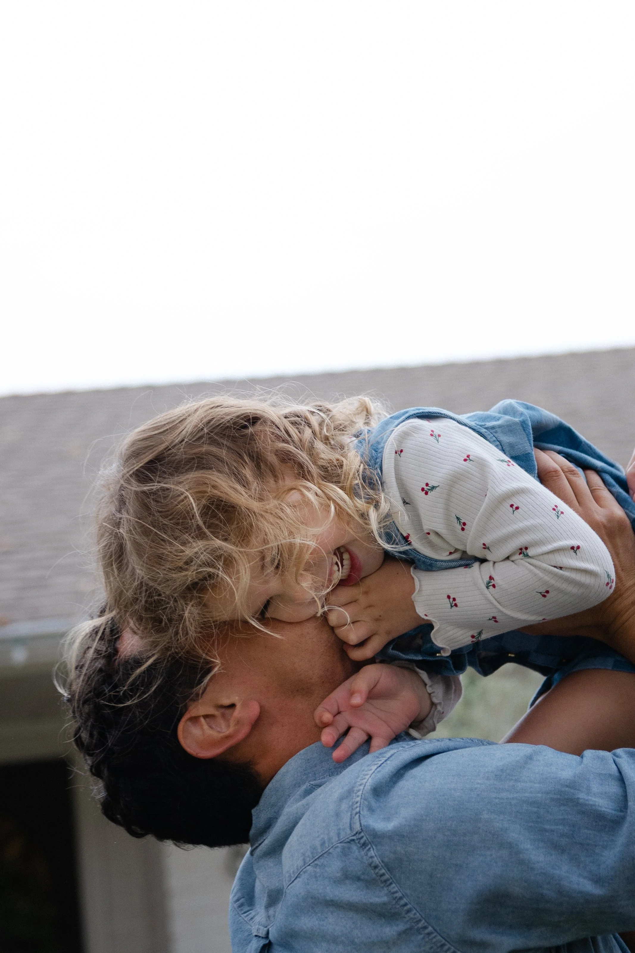 A young girl with curly blonde hair smiling and laughing while lying on a man's shoulders, with her hand on his face, outdoors in front of a house.