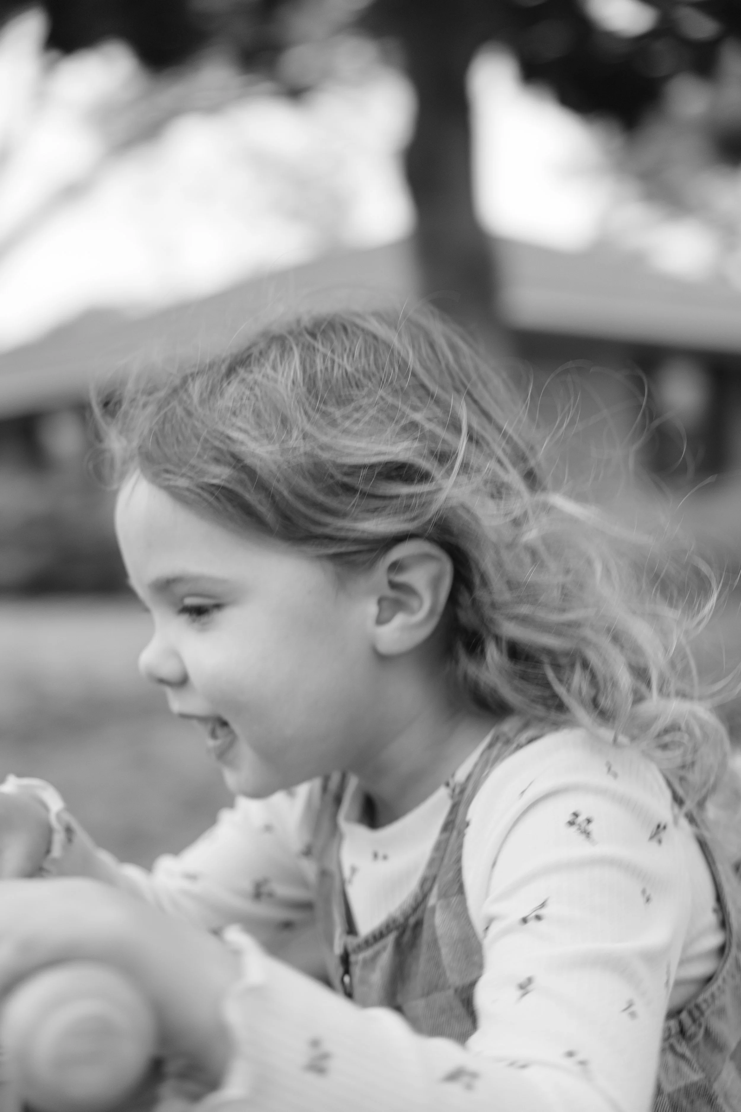 A young girl with long, wavy hair outdoors, smiling and looking to her left, wearing a long-sleeve shirt and overalls.