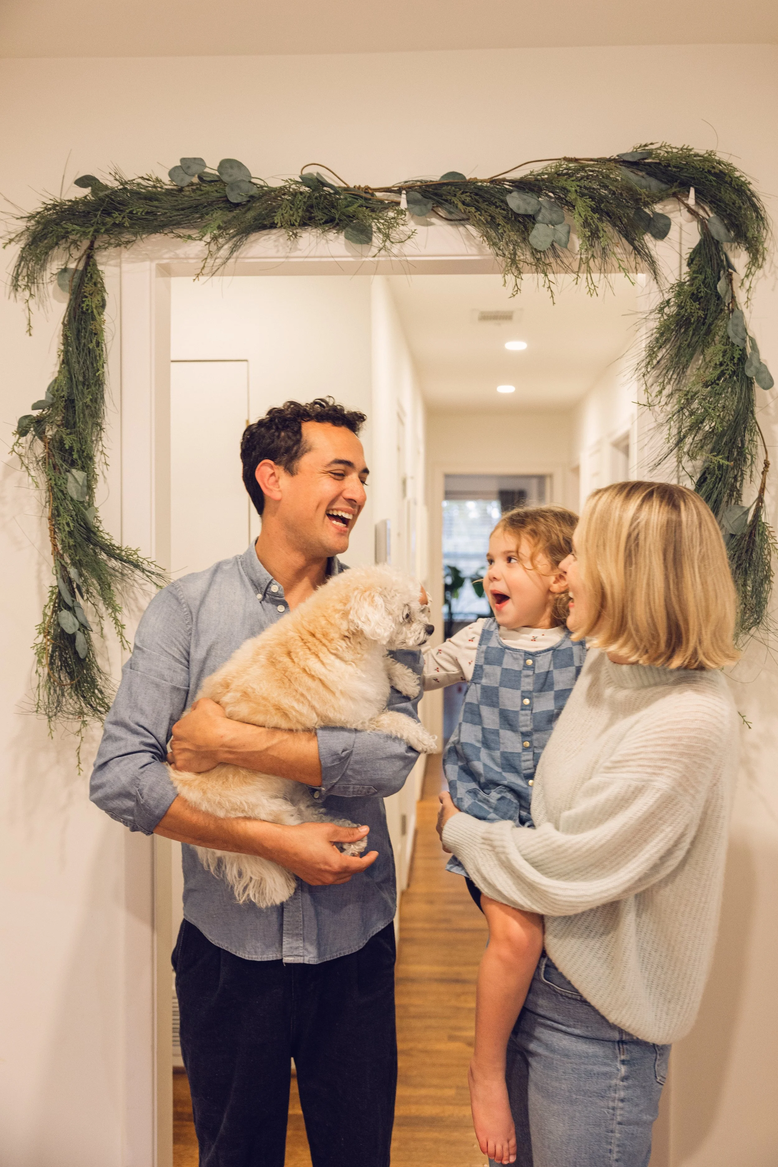 A family greeting each other at the doorway decorated with greenery, as a man holds a dog and two children and a woman smile and talk.