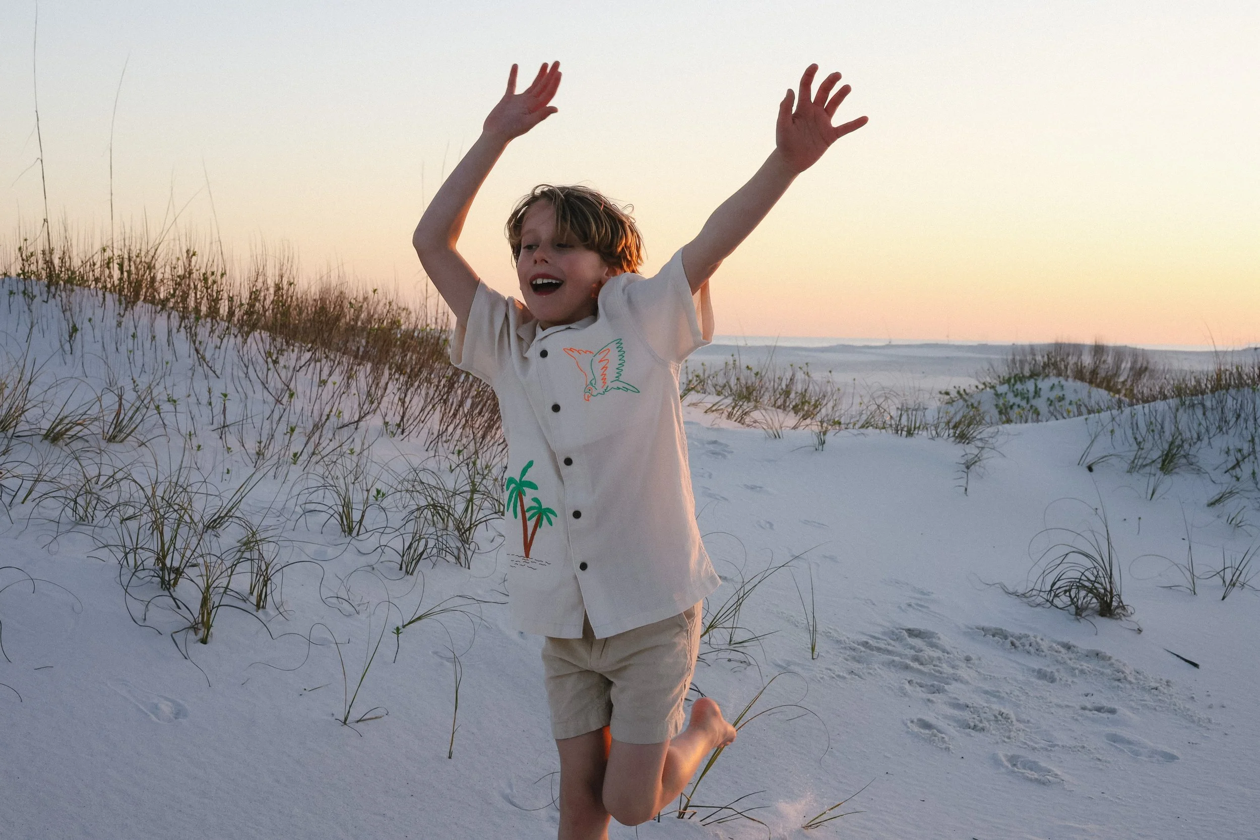 A young boy playing and jumping on sandy beach dunes at sunset, wearing a cream shirt with embroidered palm trees and birds.