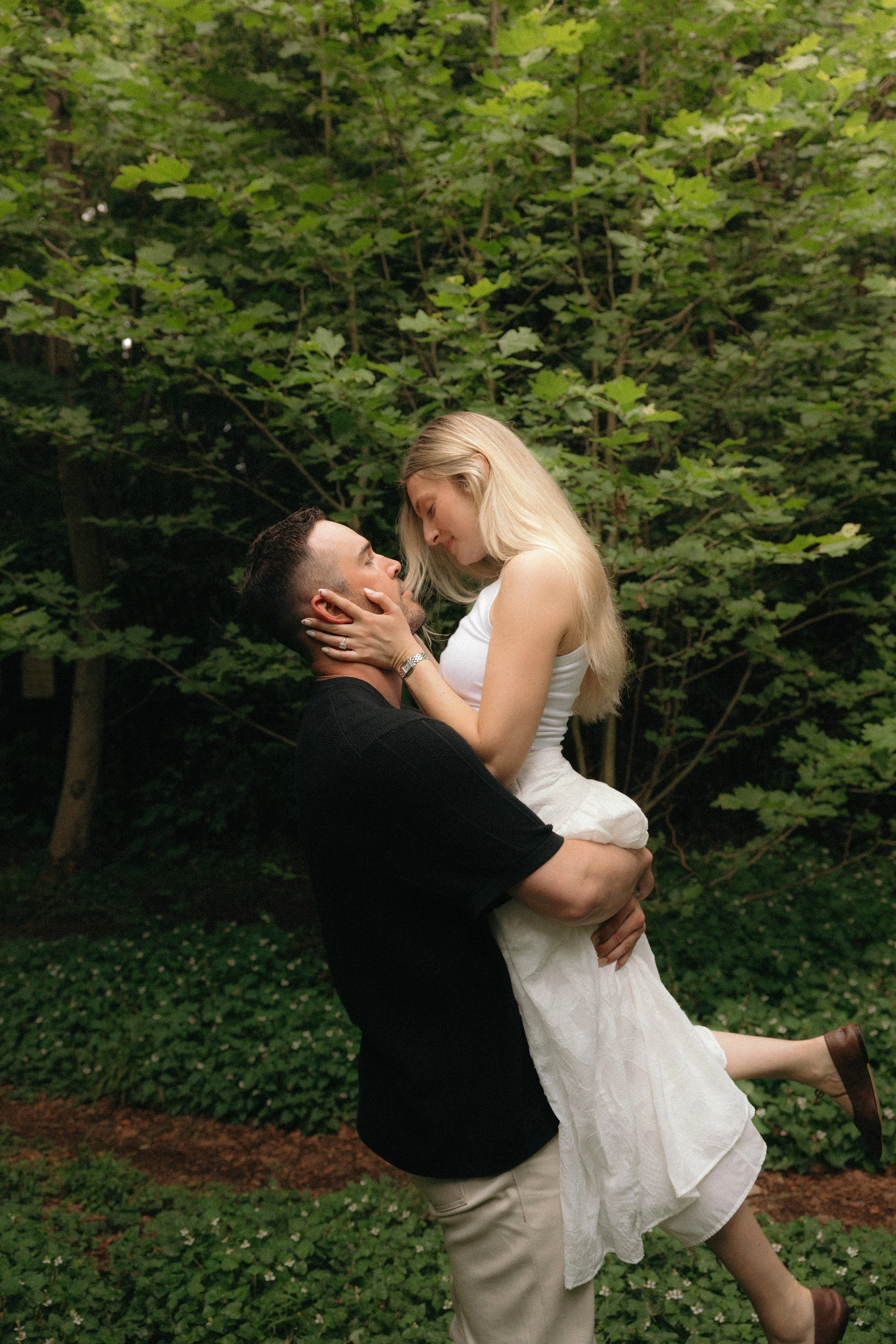 A man lifts a woman in his arms outdoors, both gazing at each other affectionately, with lush greenery in the background.