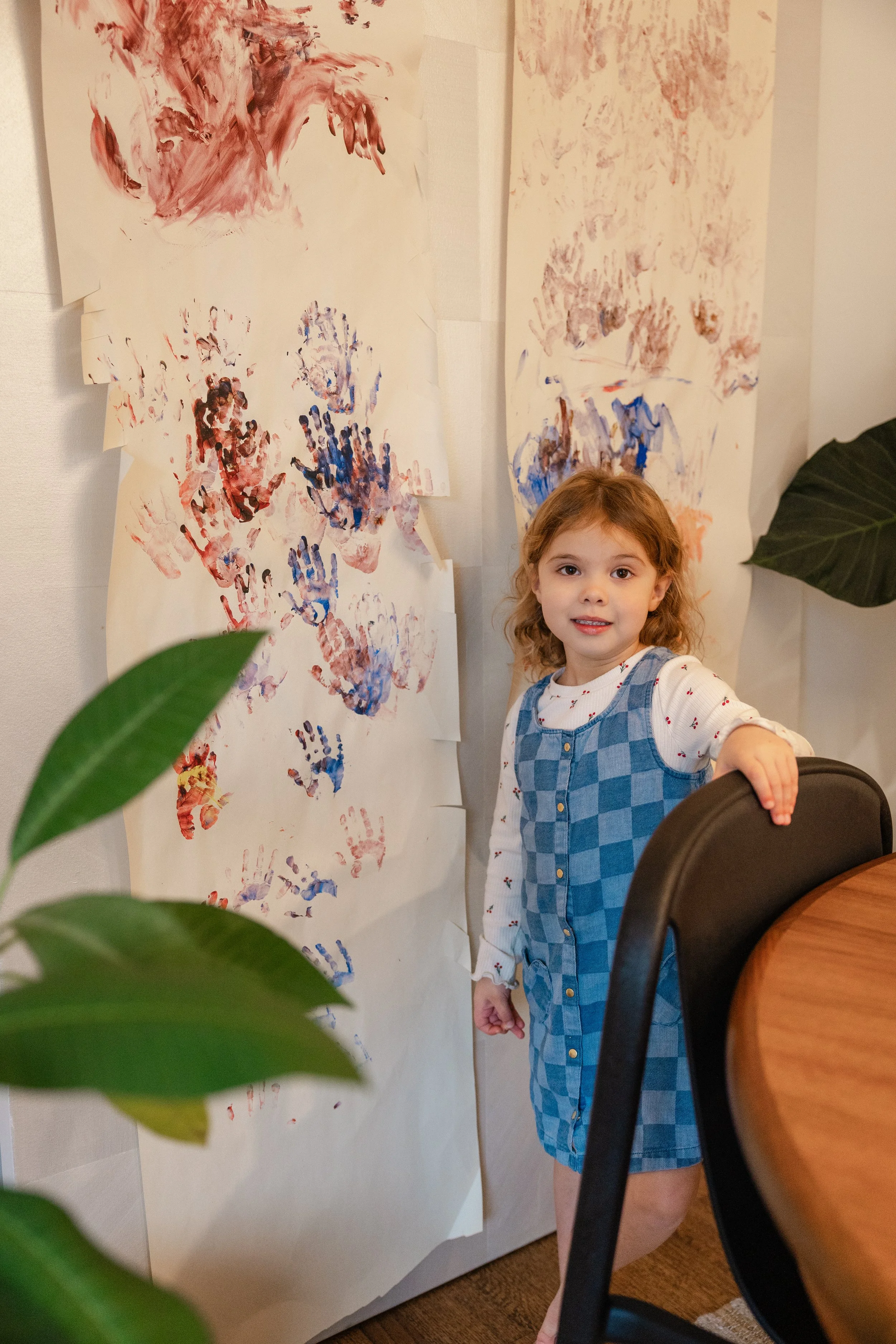 A young girl with curly hair in a blue dress standing next to a wall covered with artwork made from painted handprints.