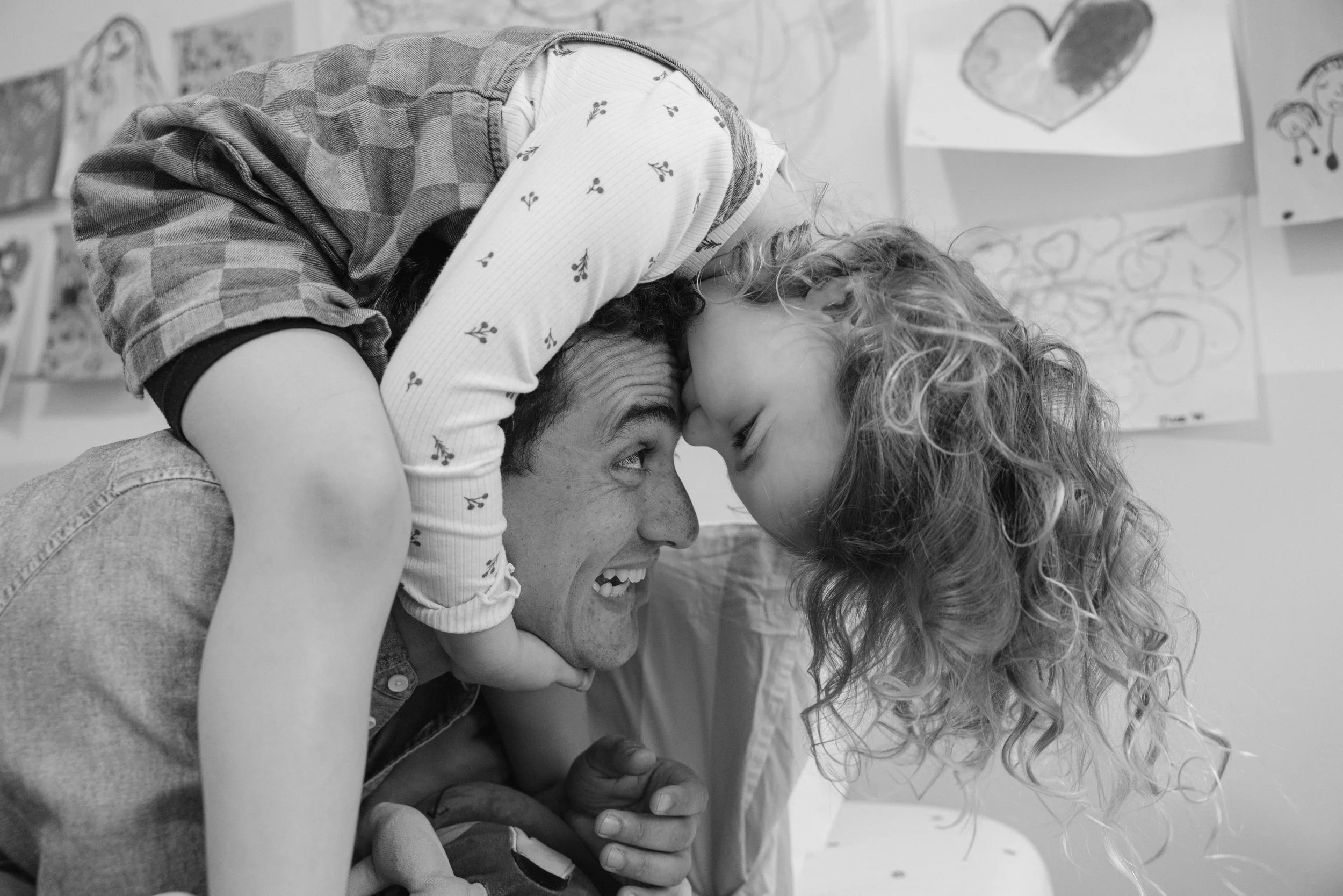 A man with curly hair and a woman with wavy hair playfully touching foreheads with a young girl, all with smiling faces, in front of children’s drawings on the wall.