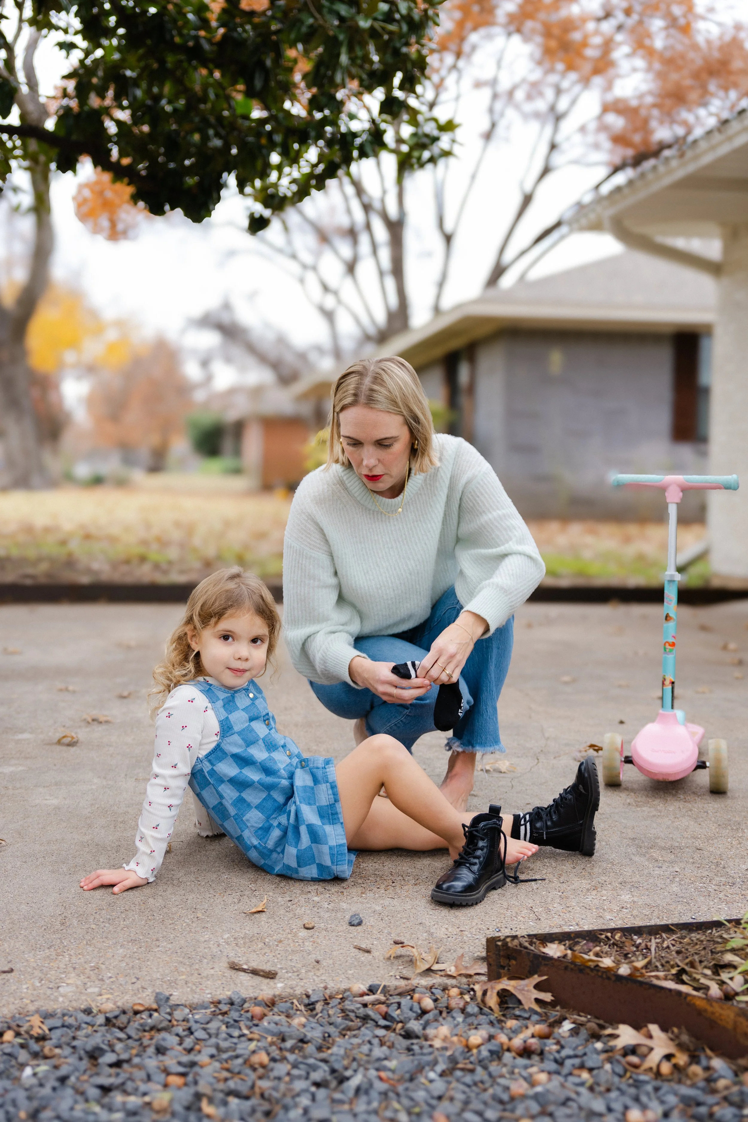 A woman and a young girl are sitting on the sidewalk in an outdoor setting. The woman is putting on a black boot on the girl, who is sitting with one leg extended. There is a pink scooter nearby. The background features houses and trees with fall foliage.