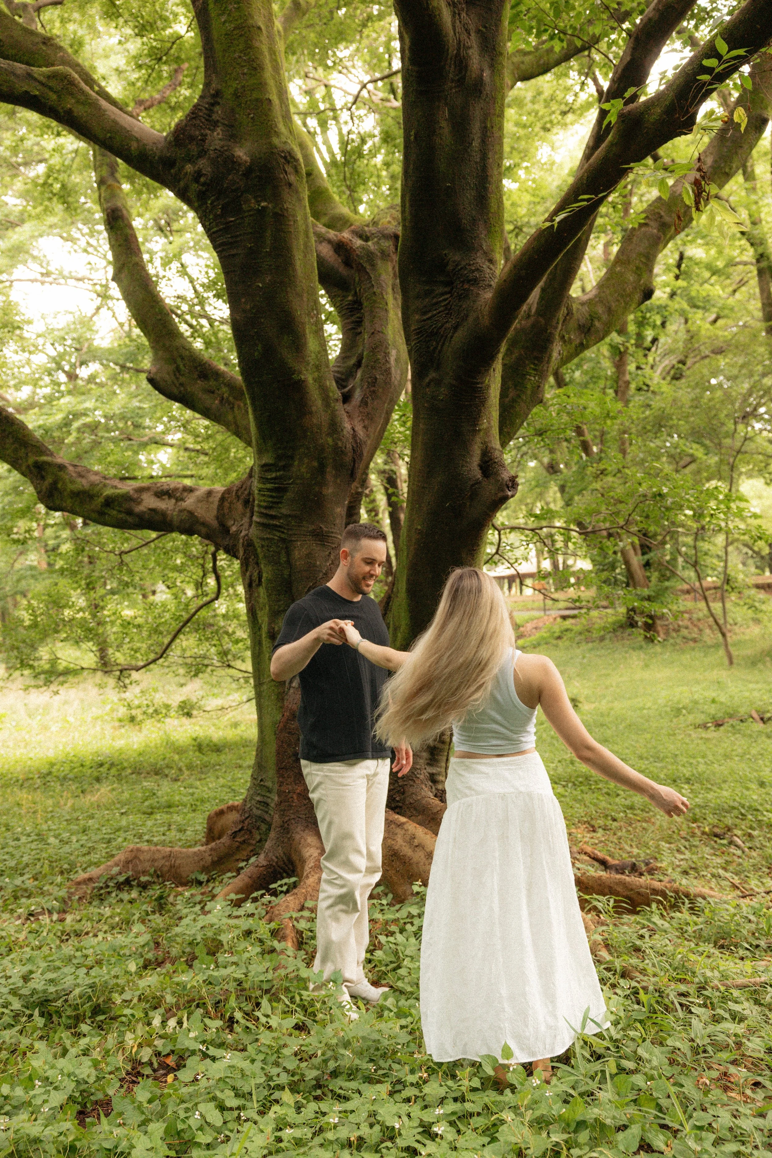 A young man and woman dancing outdoors near a large tree in a park with green foliage.