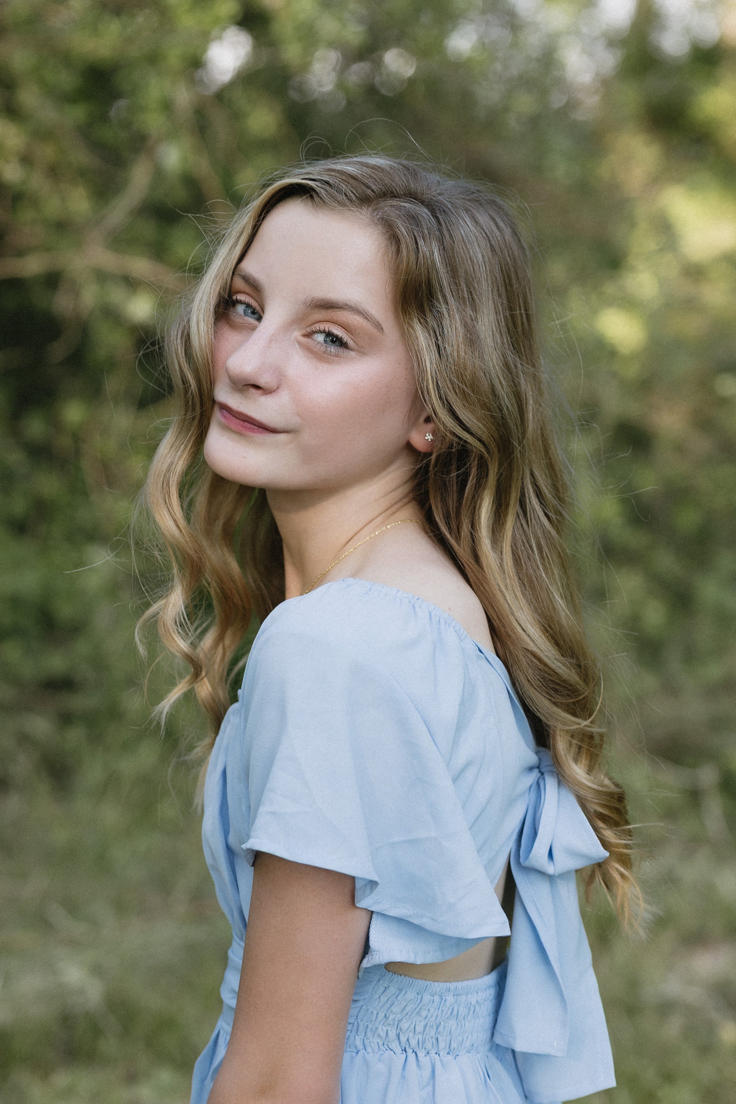 A young woman with long, wavy blonde hair standing outdoors in a natural setting with green foliage, wearing a light blue dress with puff sleeves and a bow at the back, smiling softly at the camera.
