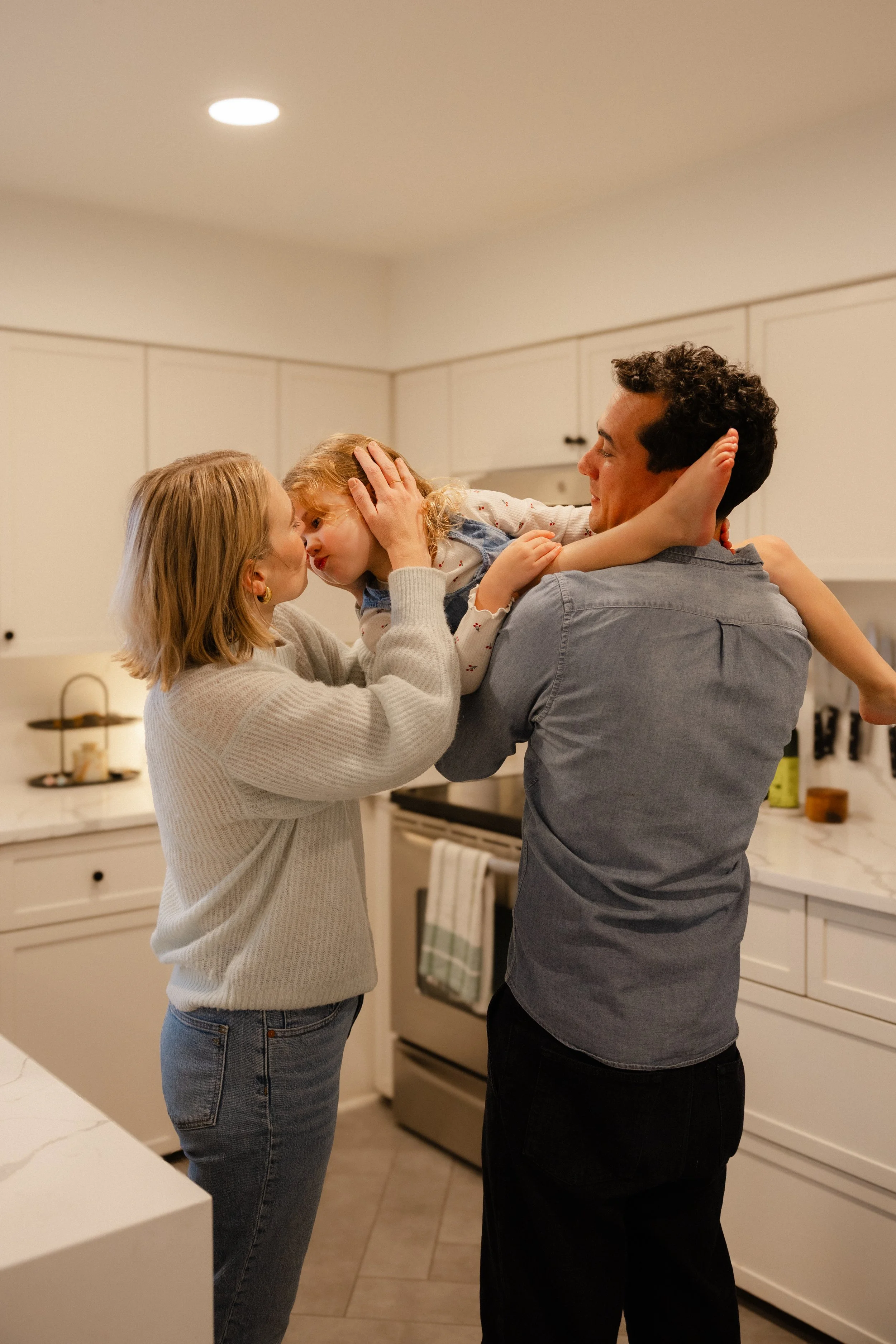 A family in a kitchen, with a woman and man holding a young girl between them, touching foreheads as they smile.
