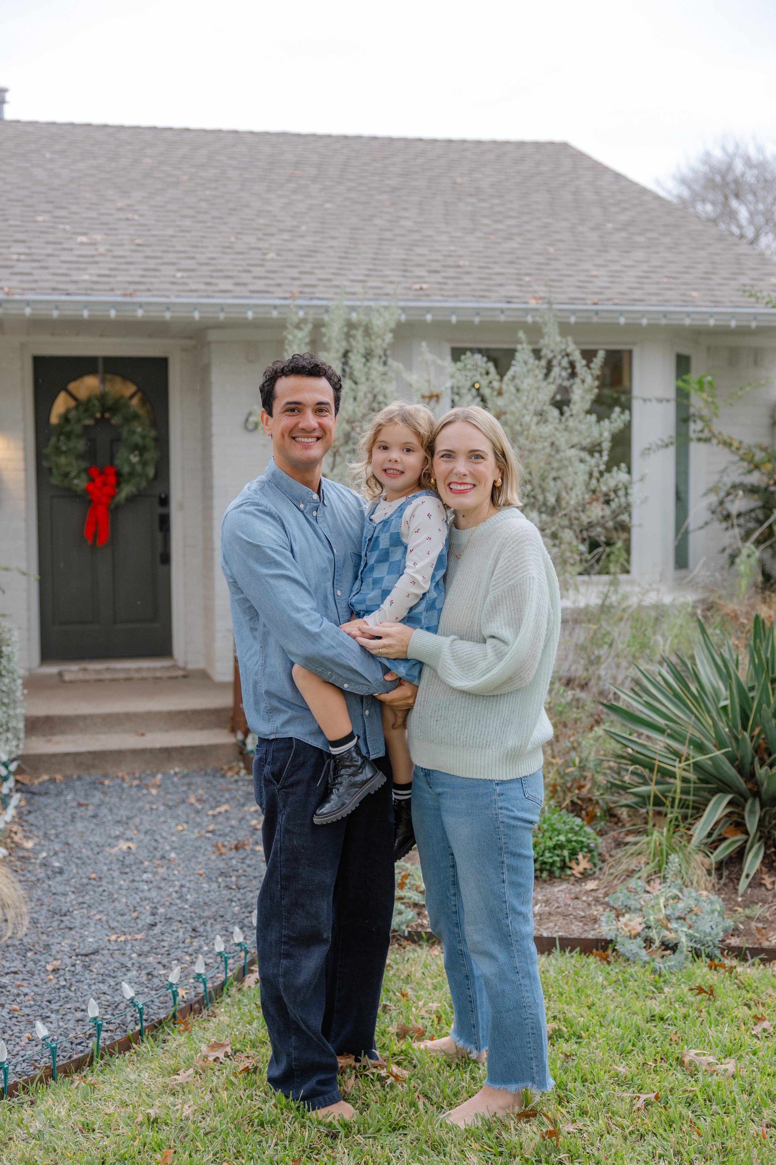 A family of three standing outside their house in front of a festive front door with Christmas decorations, smiling and holding each other on a lawn.