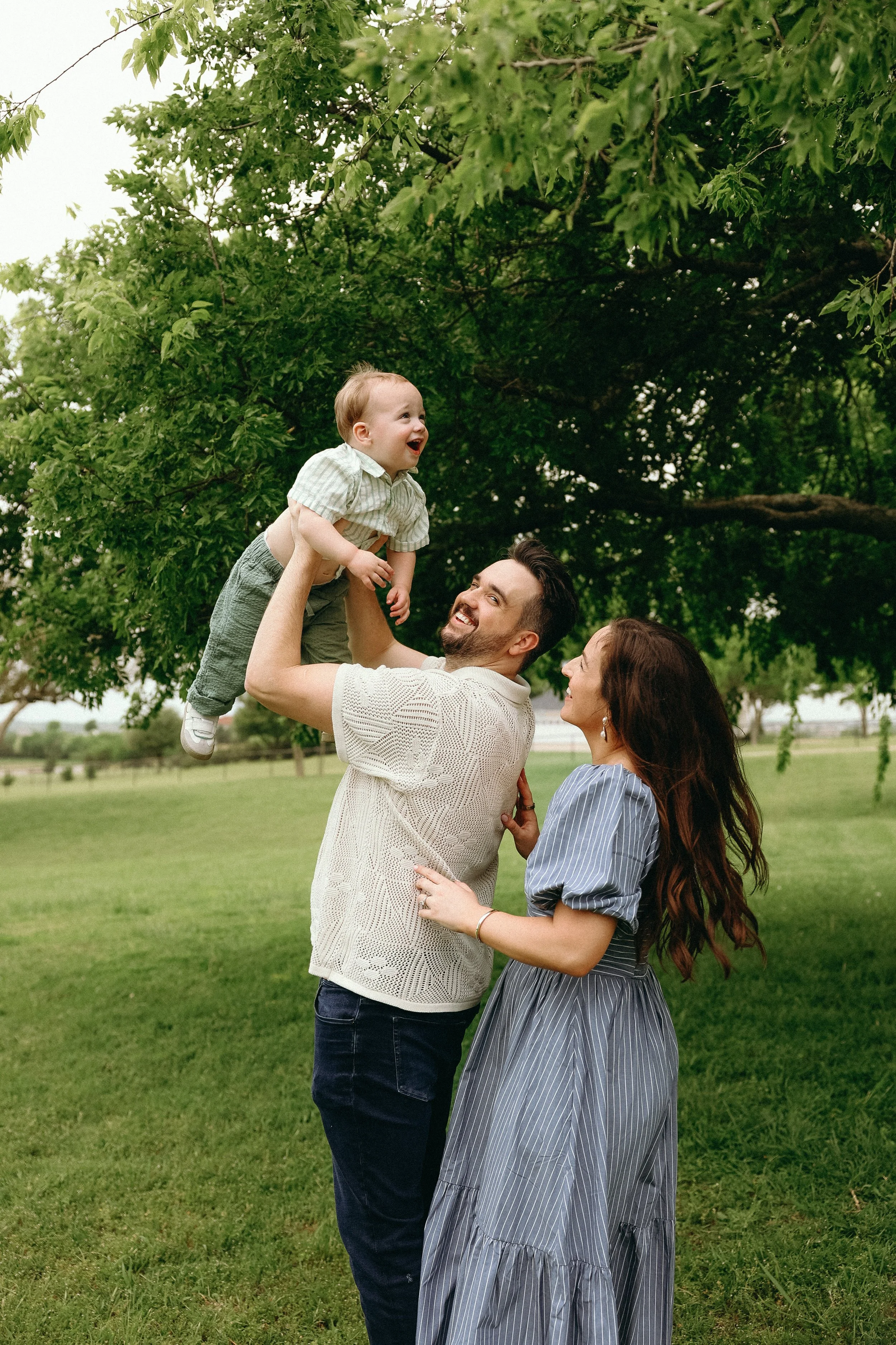 A happy family of three outdoors under a large green tree; a man lifts a young boy, who is laughing and reaching out, while a woman looks on, smiling.