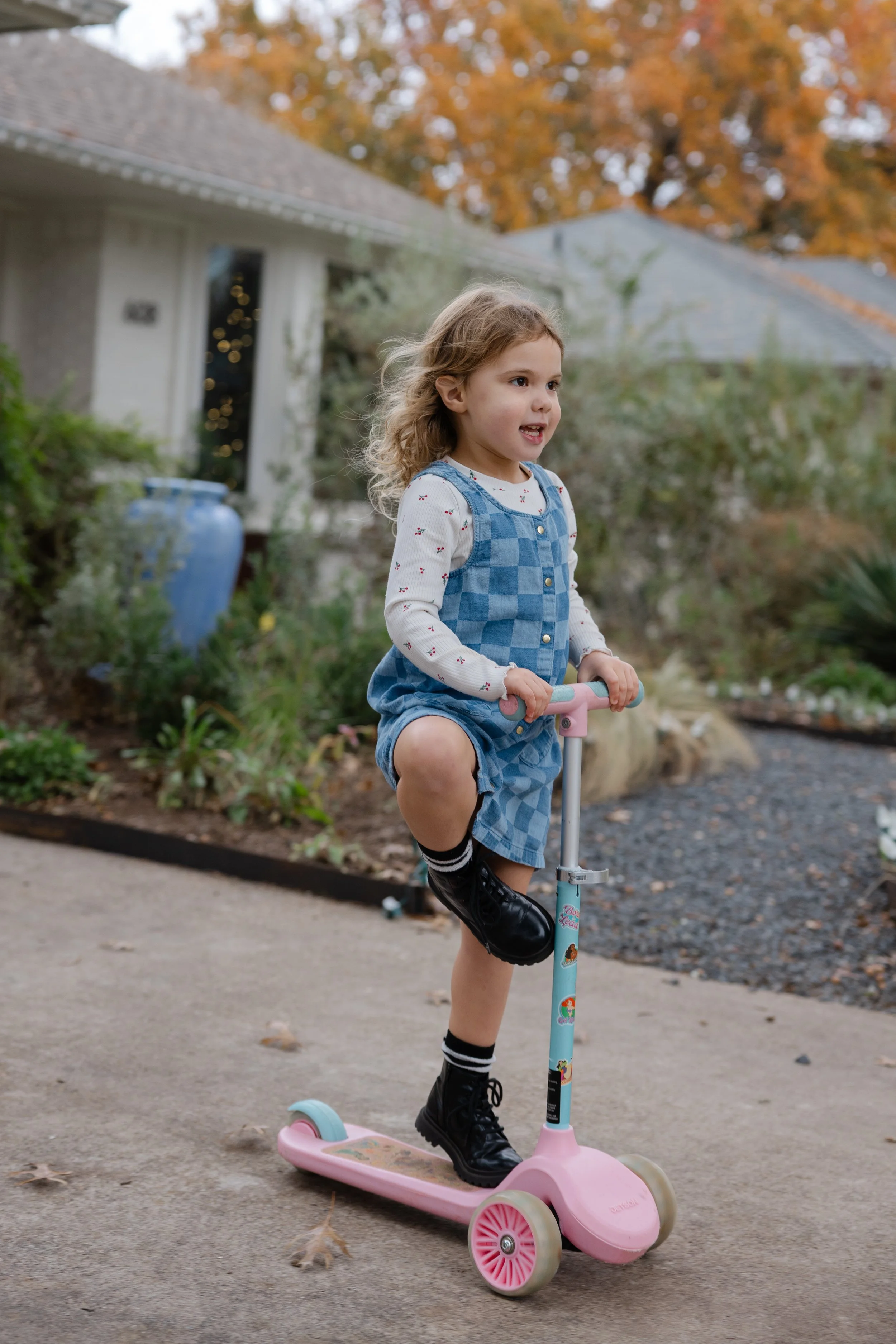 Young girl riding a pink scooter on a sidewalk outside a house with autumn-colored trees in the background.