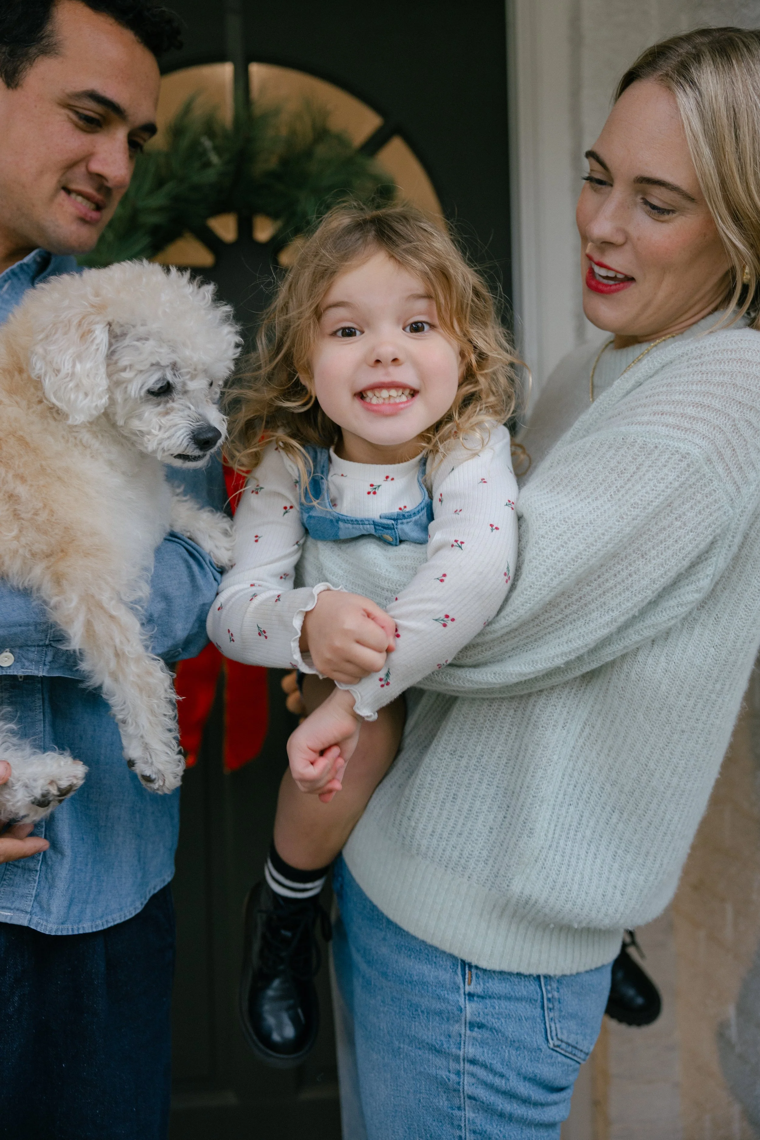 A smiling young girl with curly hair is being held by a woman, while a man holds a fluffy white puppy. All are gathered at a doorway with a wreath in the background.