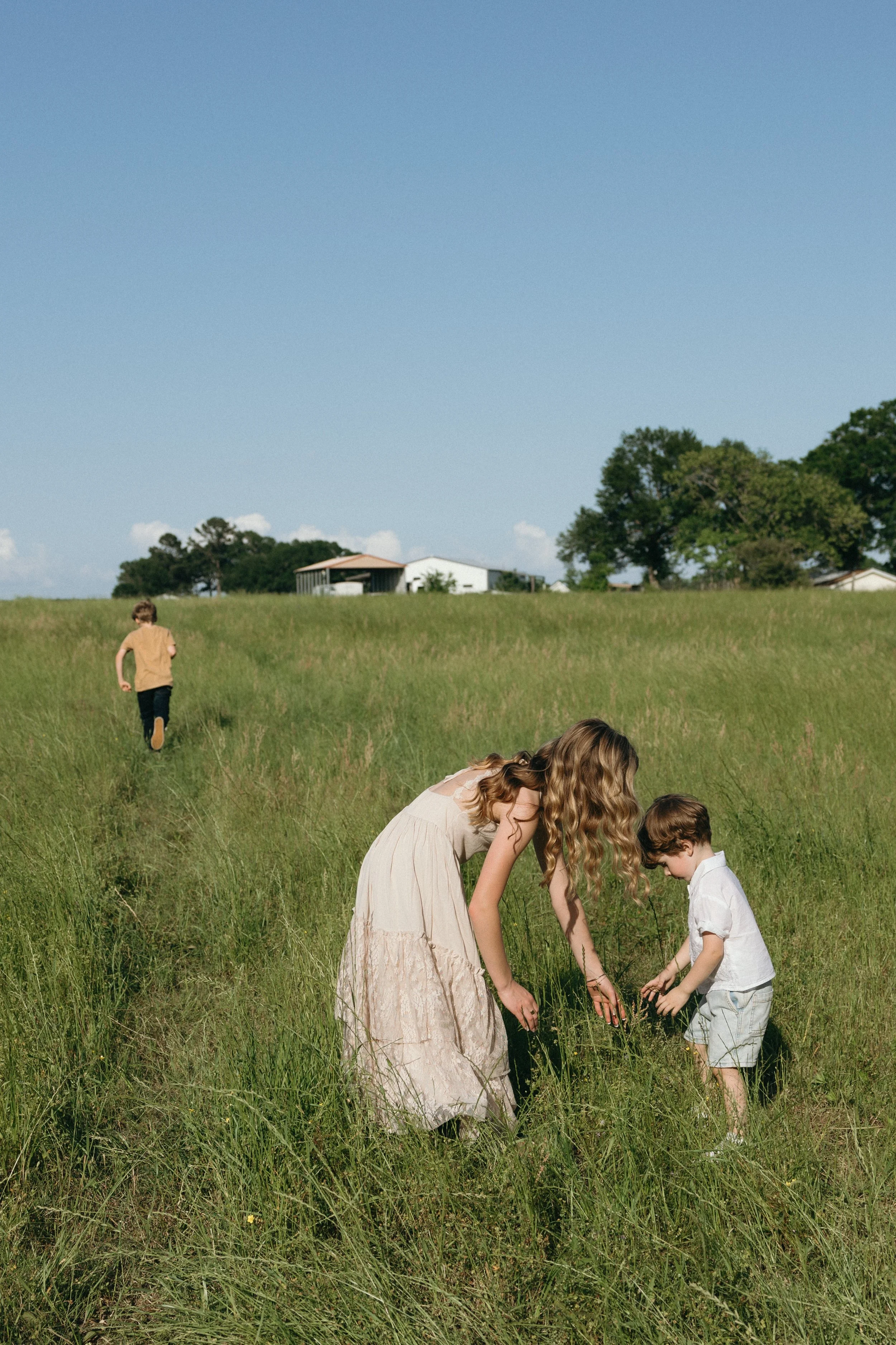 A woman with long blonde hair in a light pink dress bends down in a grassy field with two small boys, one in a white shirt and light shorts, and another in the distance wearing a tan shirt and dark pants, under a clear blue sky with a few trees and b