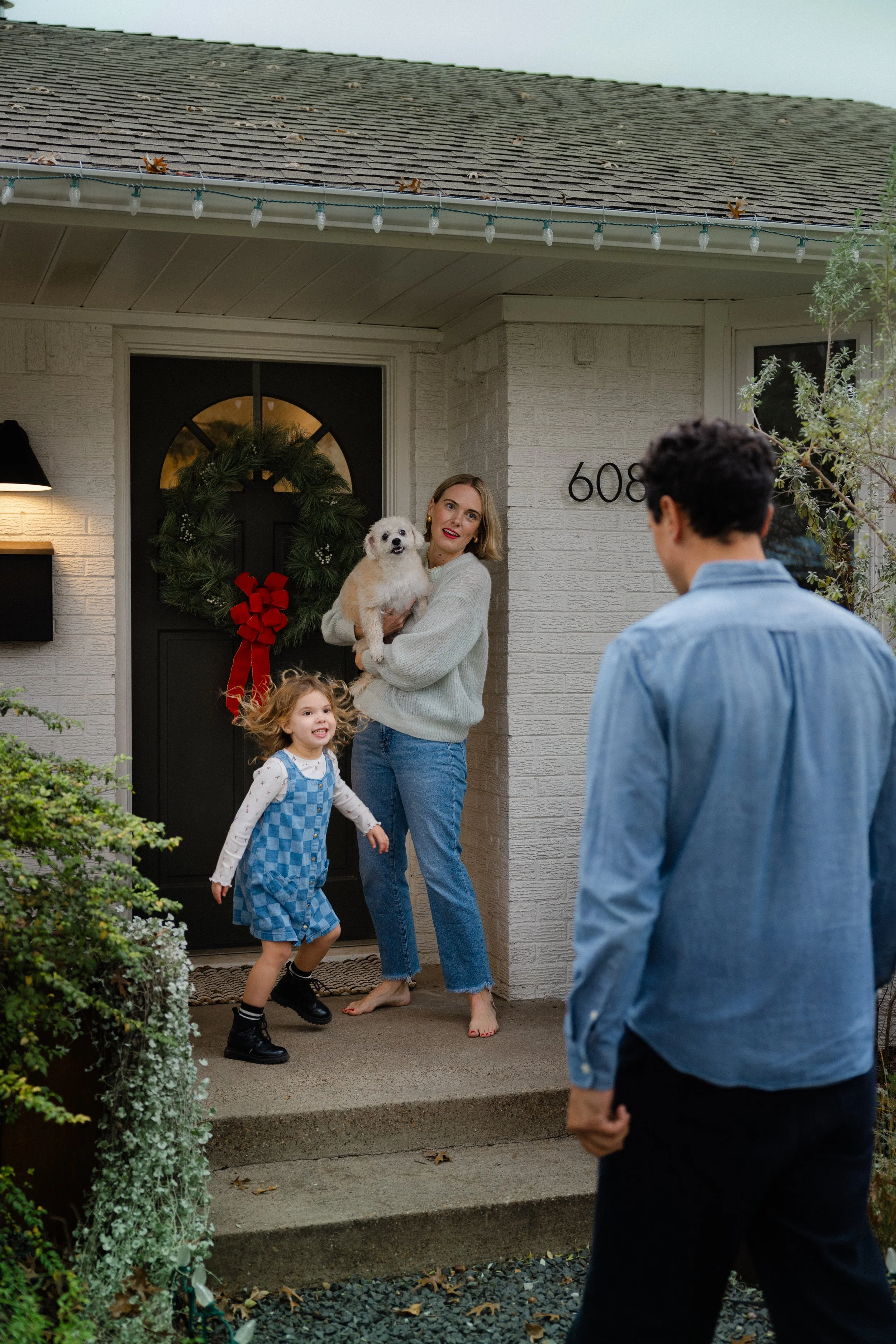 A woman holding a dog on the front porch of a house, with a young girl dancing nearby, and a man standing in the front yard, all greeting others at the door decorated for the holidays with a wreath and Christmas lights.