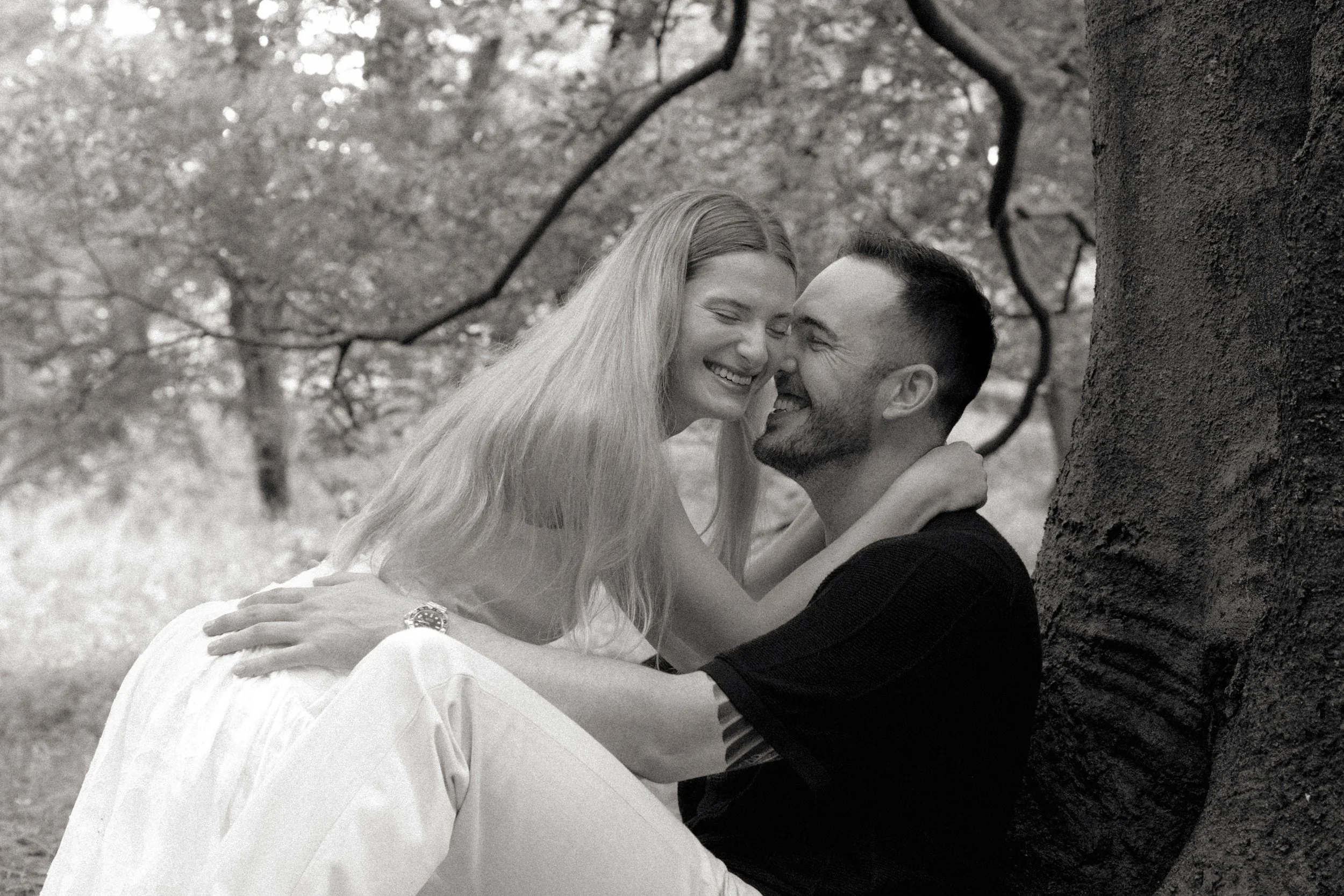 A happy couple sitting near a tree, sharing an intimate moment and smiling at each other in a park or natural setting.