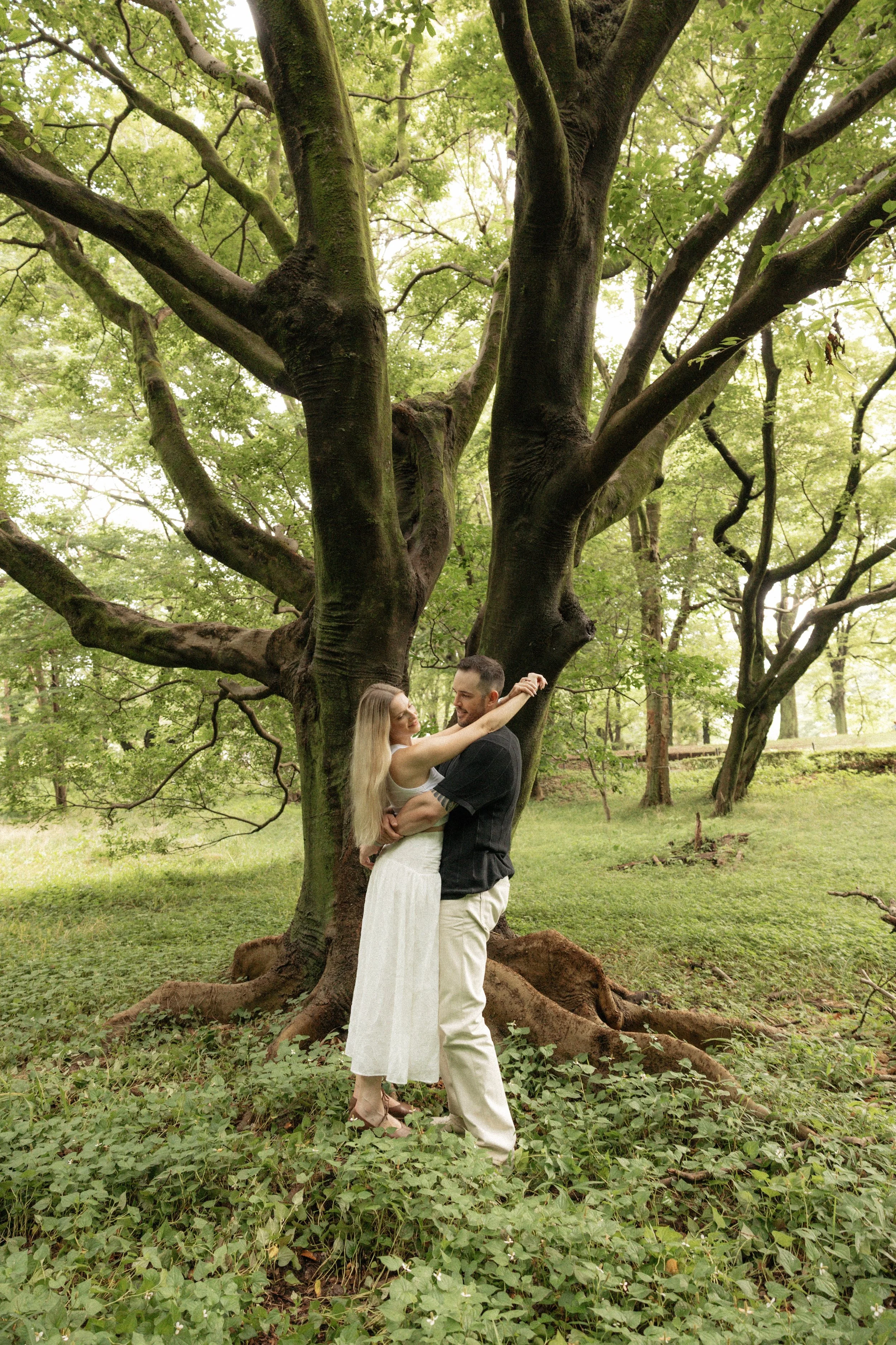 A newly engaged couple dancing under a large green tree in a lush park, with the woman wearing a white dress and the man wearing a black shirt and cream pants. engagement photo session.