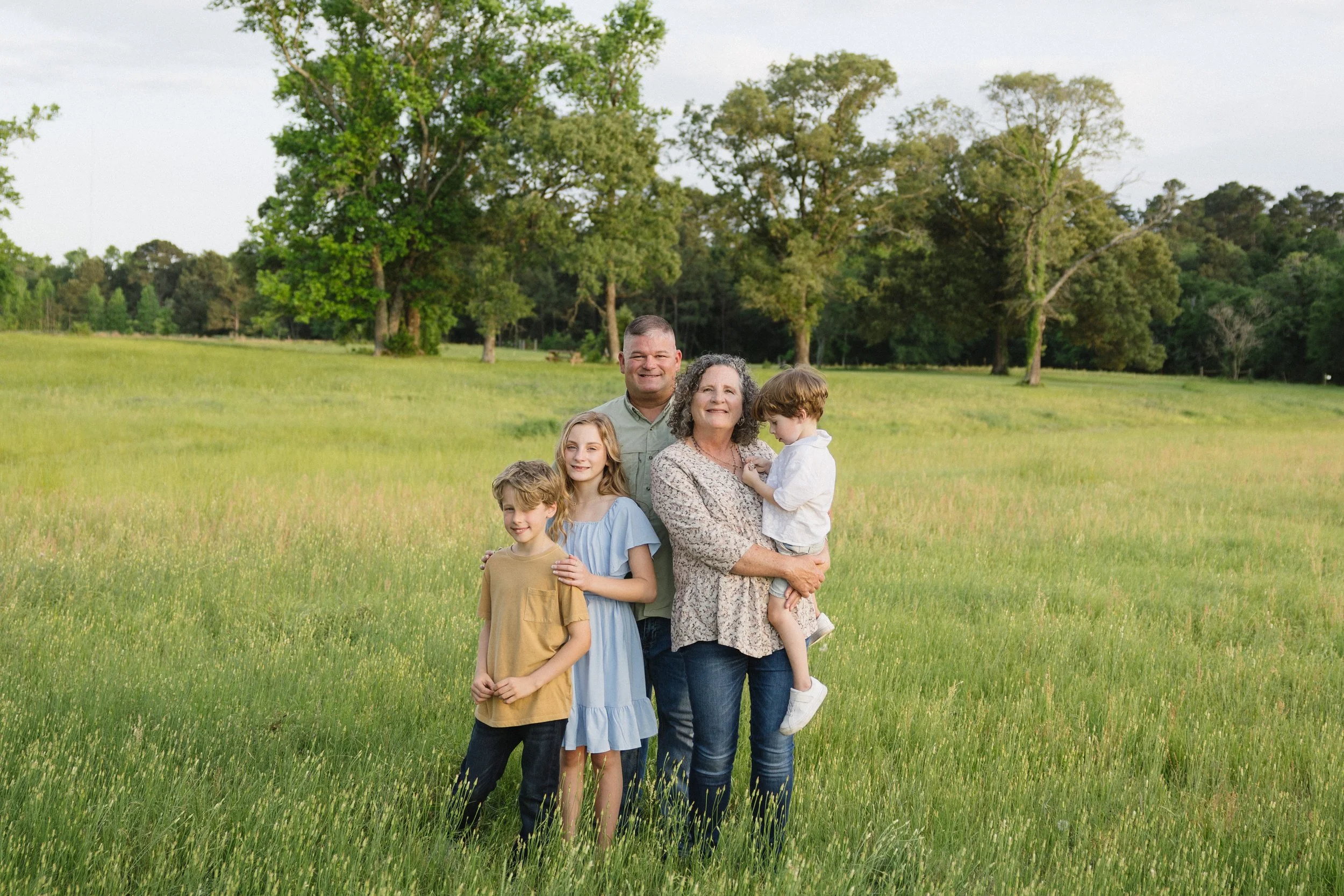 A family of five standing in a grassy field with trees in the background, smiling at the camera.