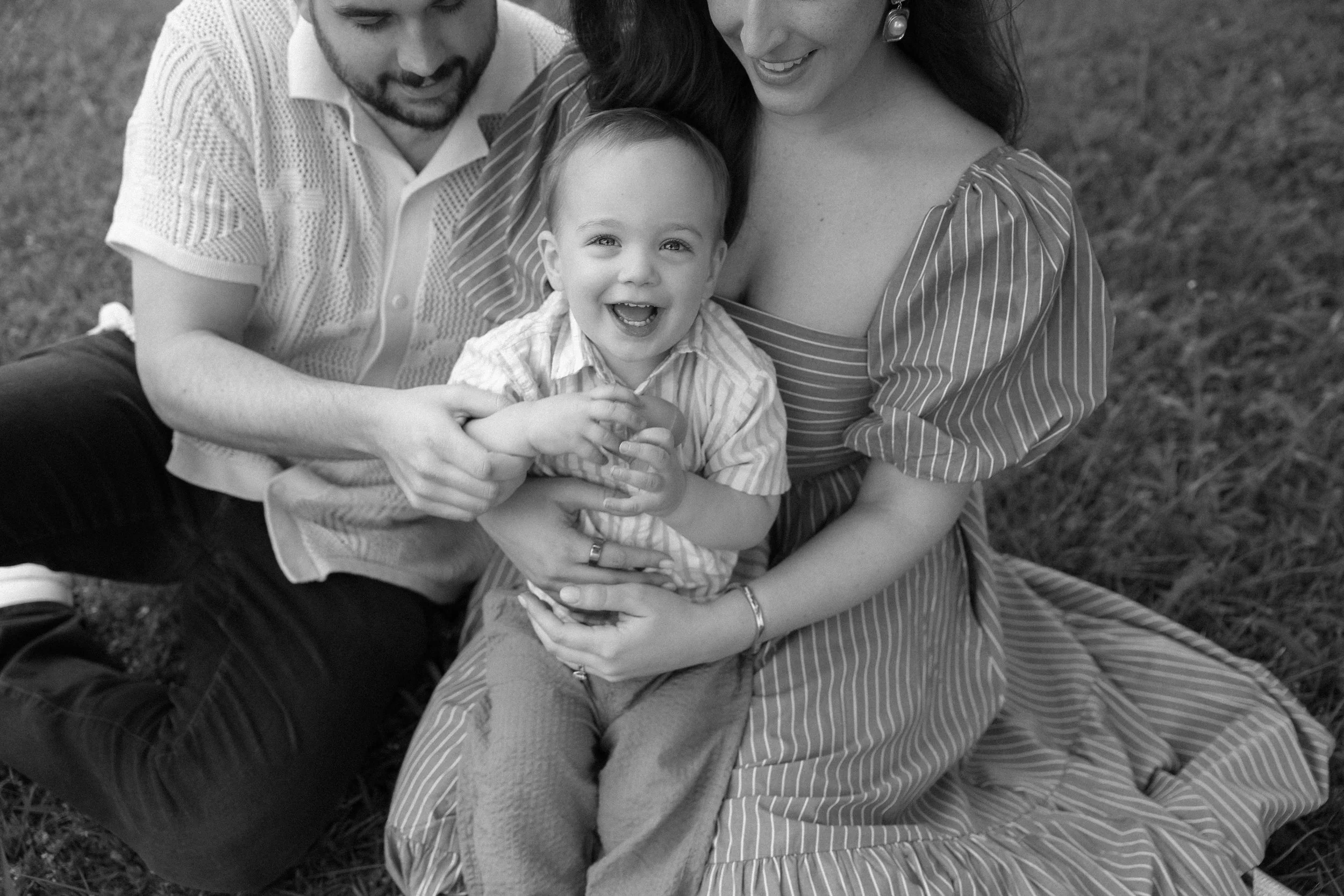 A happy family of three sitting on grass outdoors, with a smiling young boy in the middle, being embraced by a man and woman.