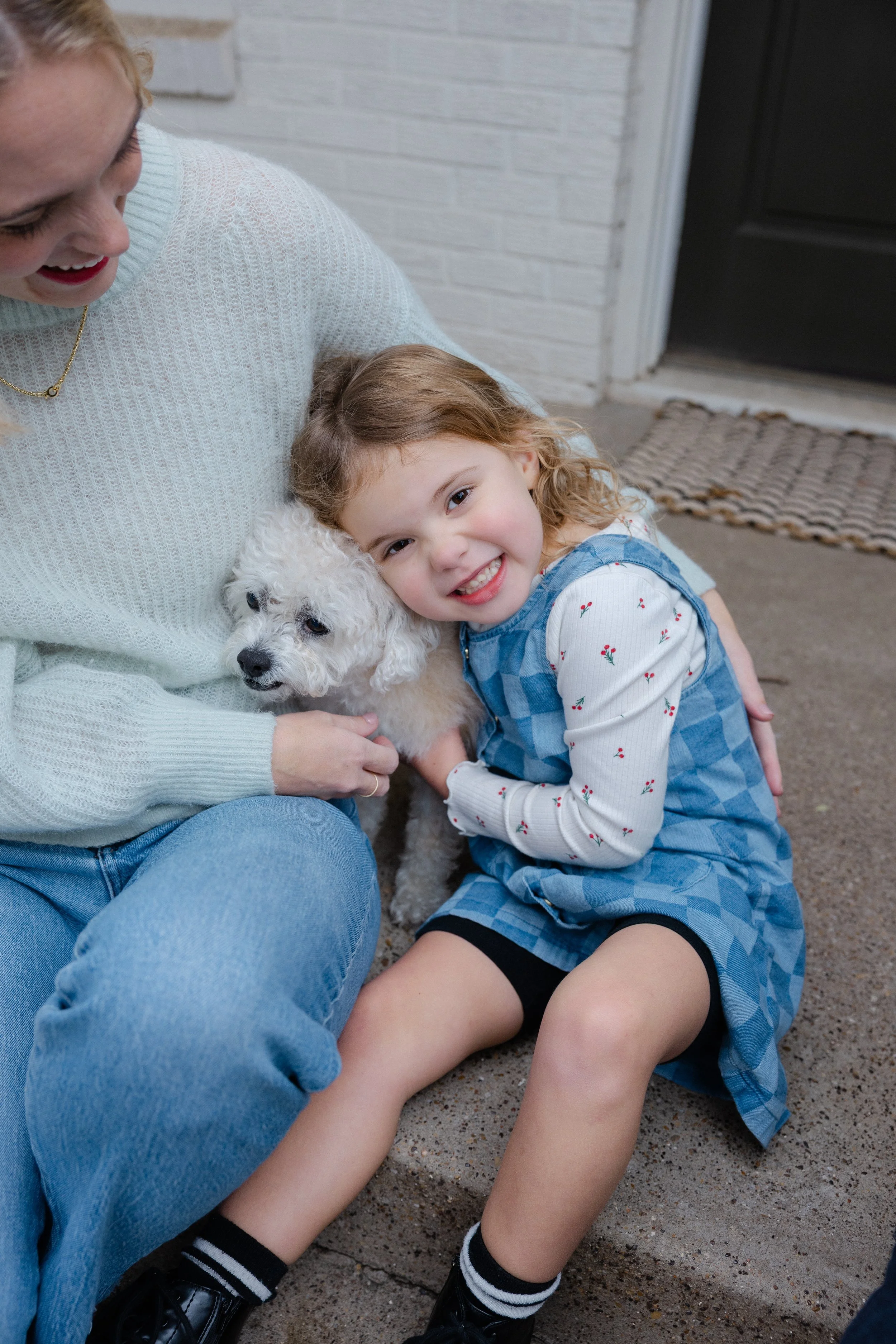 A young girl with curly hair smiling and hugging a white poodle sitting on a concrete step, with an adult woman next to her.