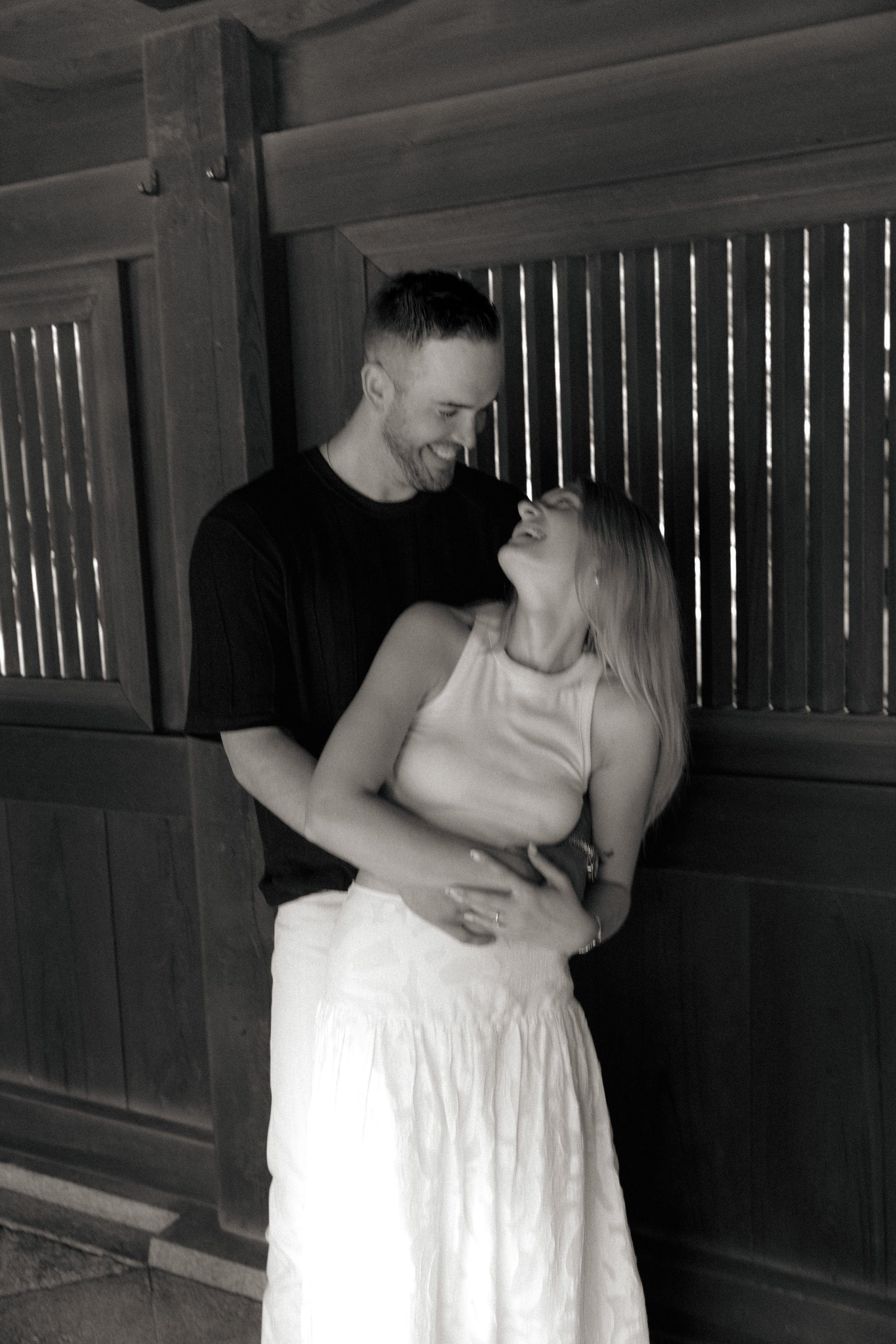 A man and woman sharing a joyful moment indoors near a wooden wall. The woman is wearing a sleeveless top and a long skirt, and the man is dressed in a dark t-shirt and light-colored pants. They are smiling at each other and holding hands.