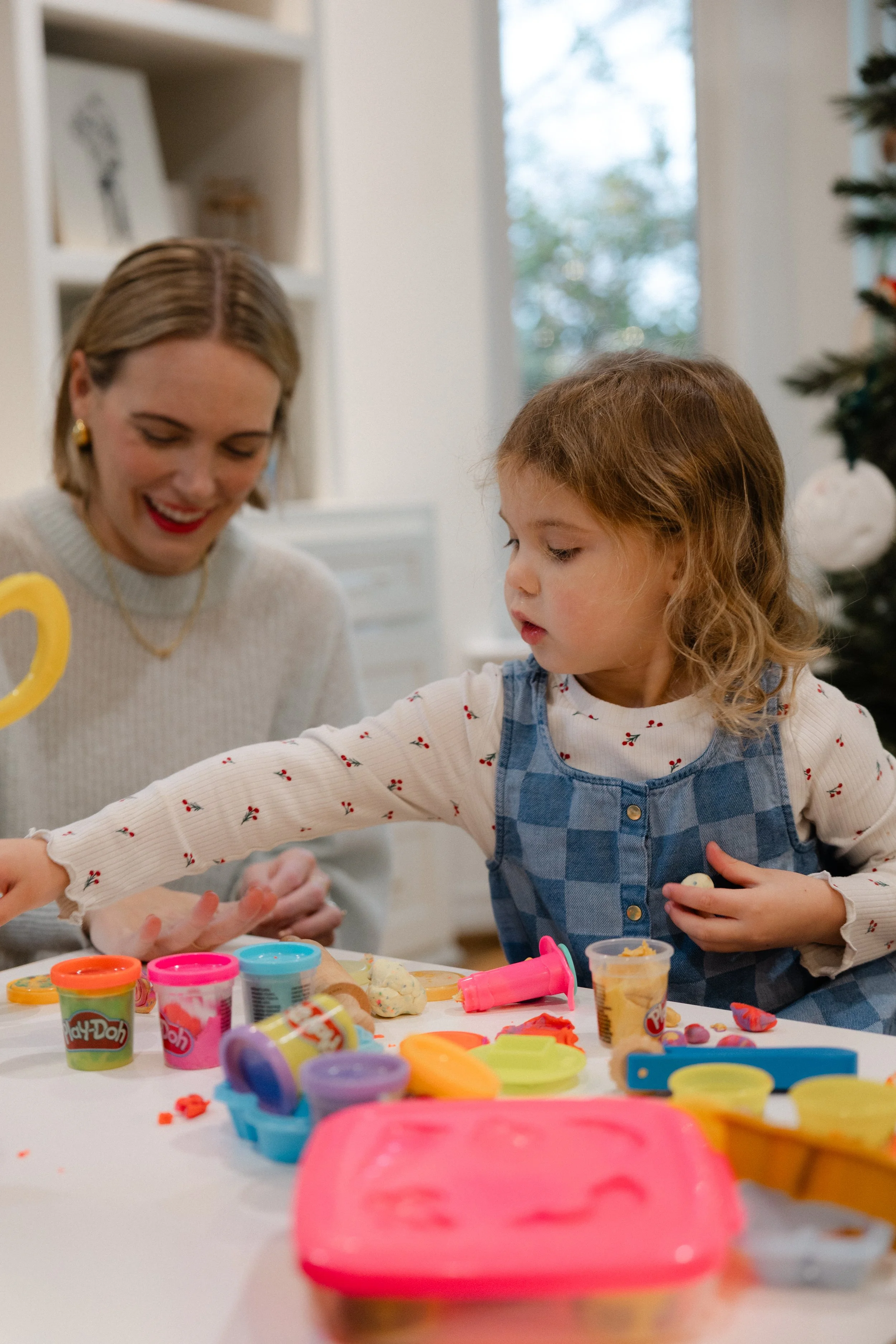 A woman and a young girl are playing with Play-Doh at a table. The girl is reaching for the Play-Doh while the woman smiles in the background. There is a decorated Christmas tree and a window in the background.