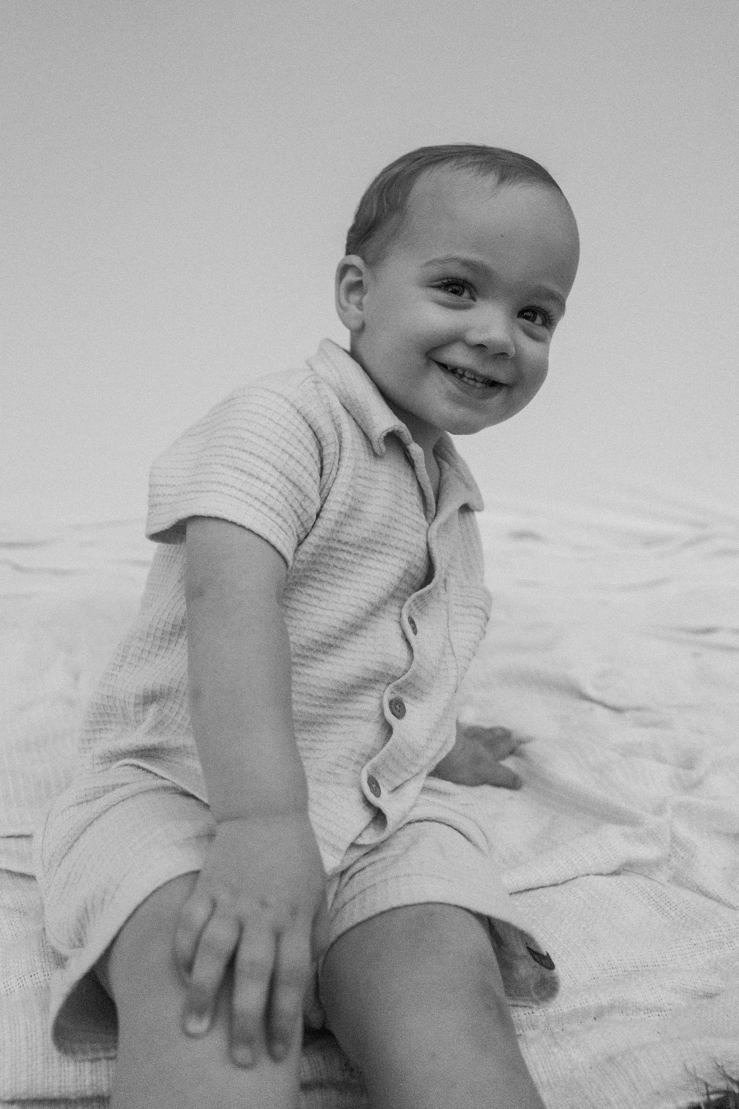 A smiling young boy in a striped short-sleeve shirt and shorts sitting on a blanket outdoors.