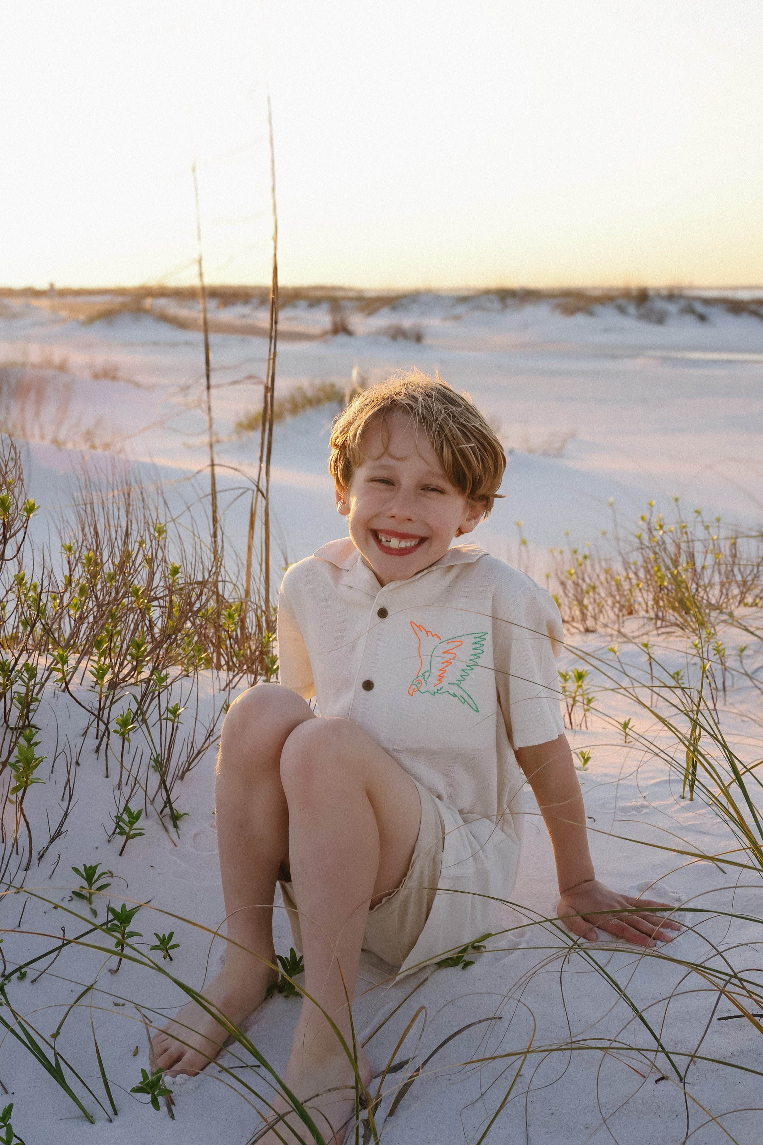 A young boy sitting on the sand at the beach, smiling with his eyes closed, surrounded by grass and dunes during sunset.