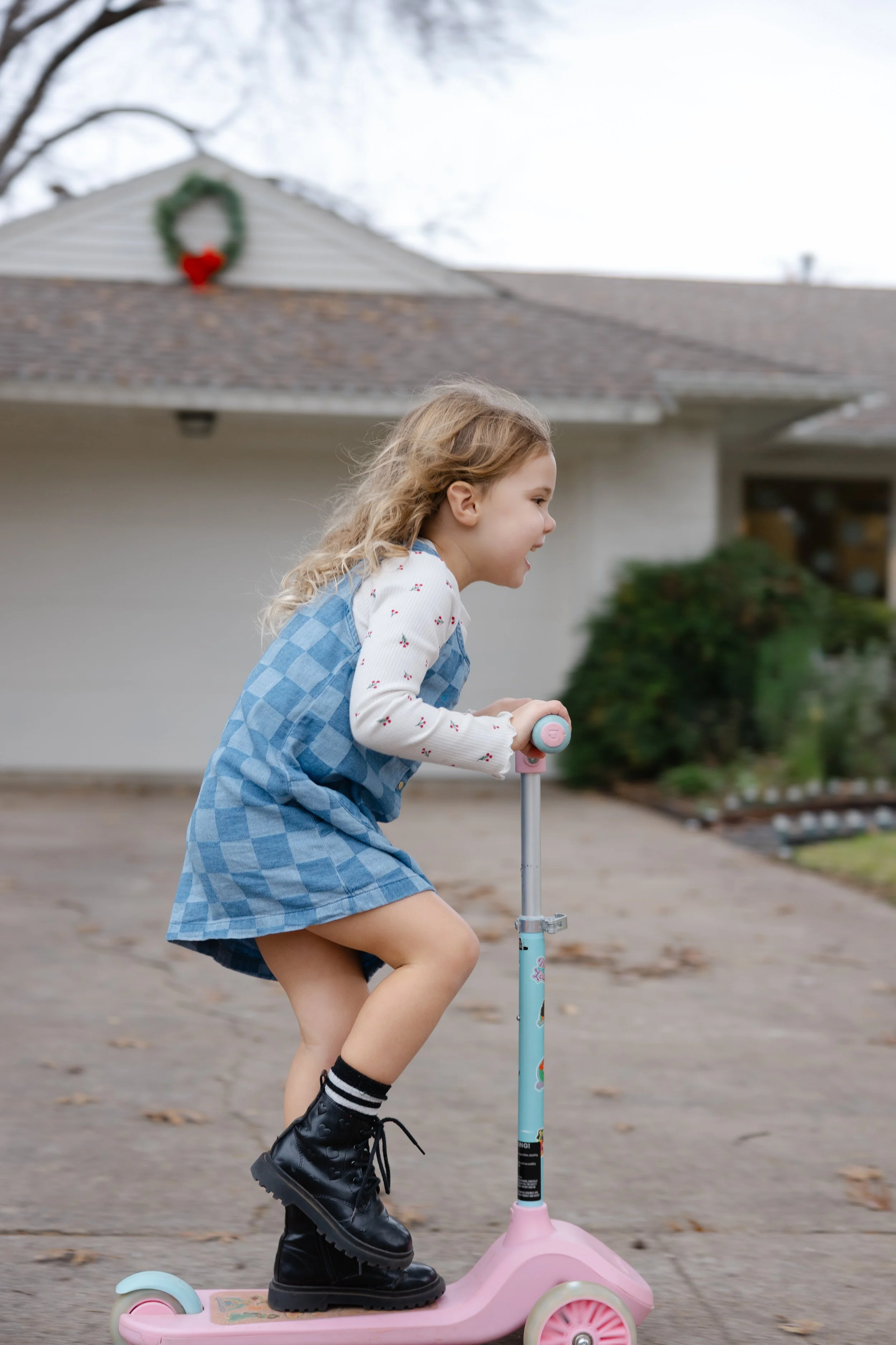 Young girl riding a pink scooter outdoors in front of a house with a wreath on the roof.